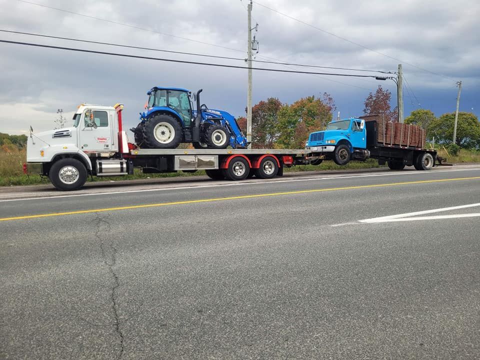 White truck hauling a blue tractor and a blue truck loaded with brown pallets on a highway. Overcast sky.