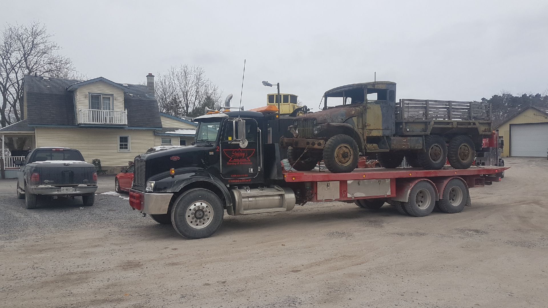 Black semi-truck hauling a vintage flatbed truck on a trailer, parked in front of a house on a cloudy day.