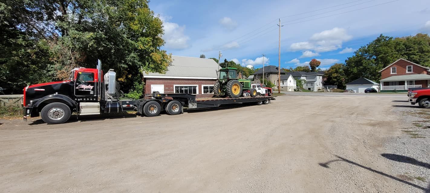 Black and red semi-truck hauling a green tractor on a flatbed trailer, parked on a gravel lot near buildings and trees.