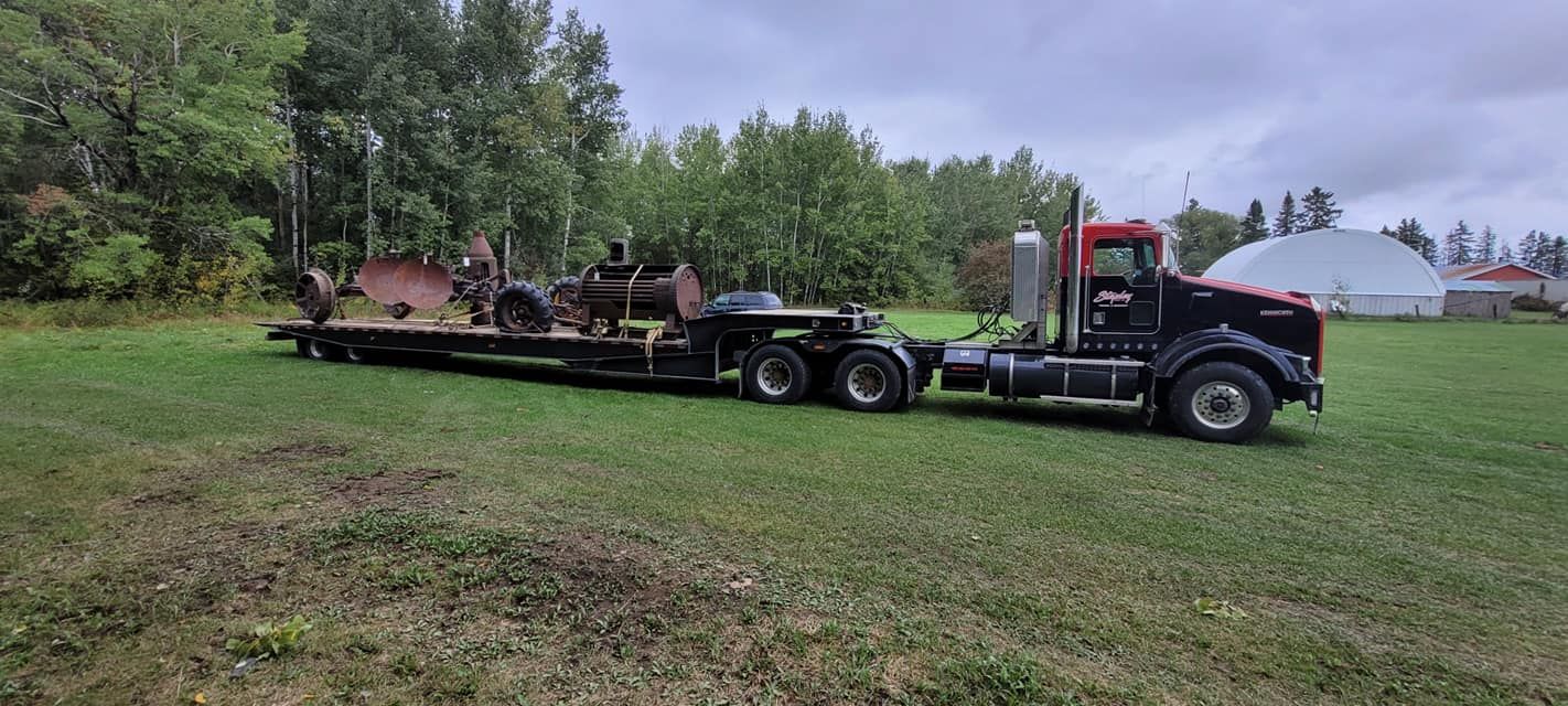 A semi-truck hauling equipment on a flatbed trailer in a grassy field. Overcast sky, trees in the background.