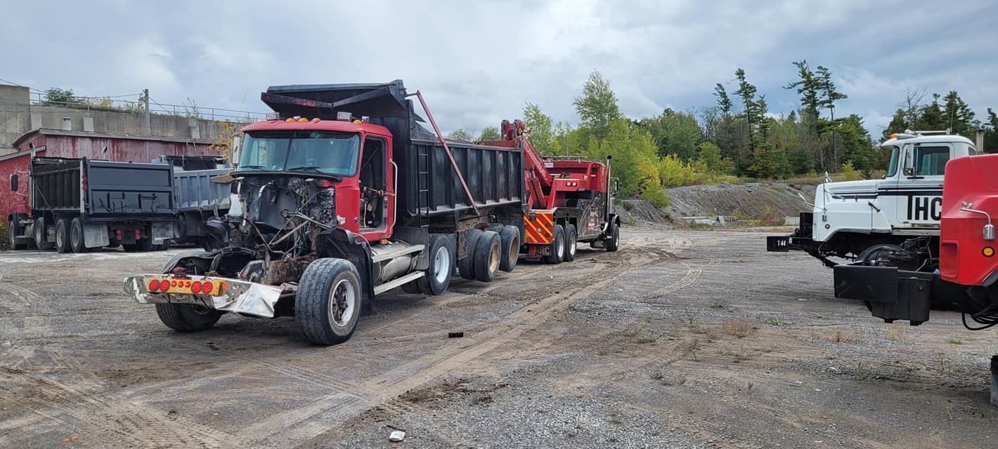 Red dump truck with missing front end parked in a gravel lot with other construction equipment.
