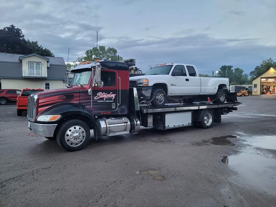 Tow truck with a white pickup truck loaded on its flatbed.  Gray sky, asphalt parking lot. 