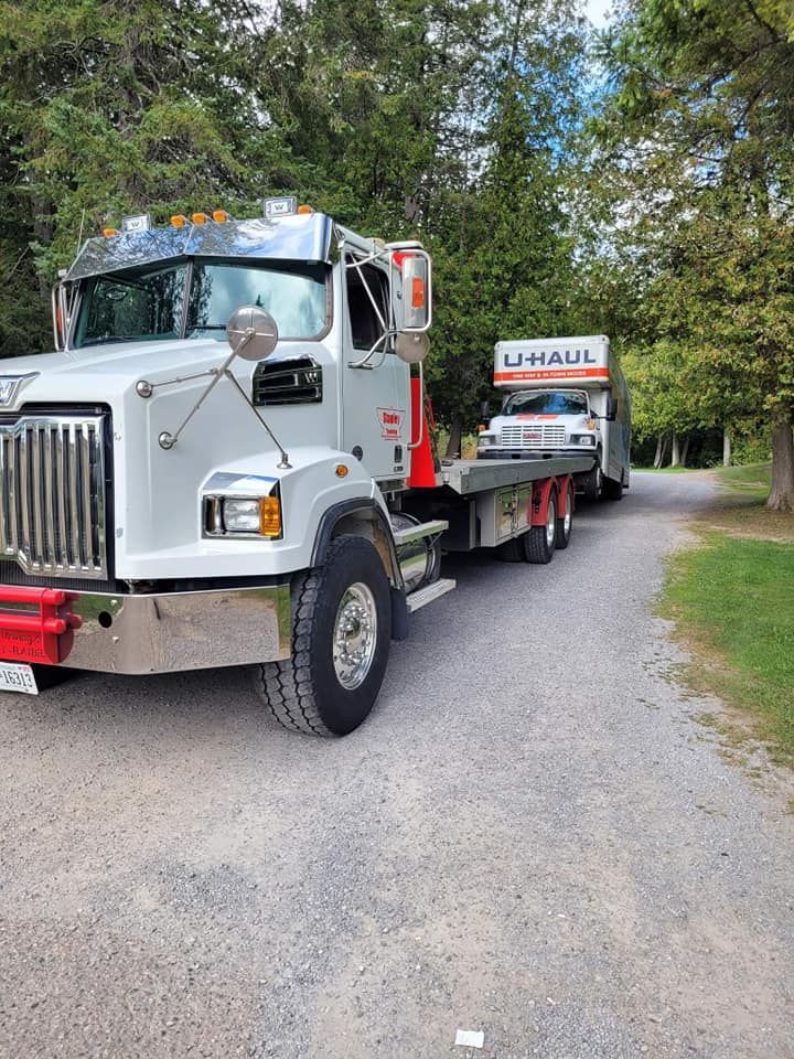 White semi-truck hauling a U-Haul trailer on a gravel road, with trees in the background.