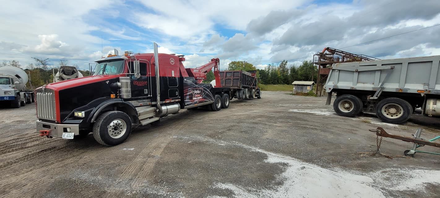 A red and black semi-truck with a trailer hauling a load of gravel. Trucks parked on a gravel lot under cloudy skies.
