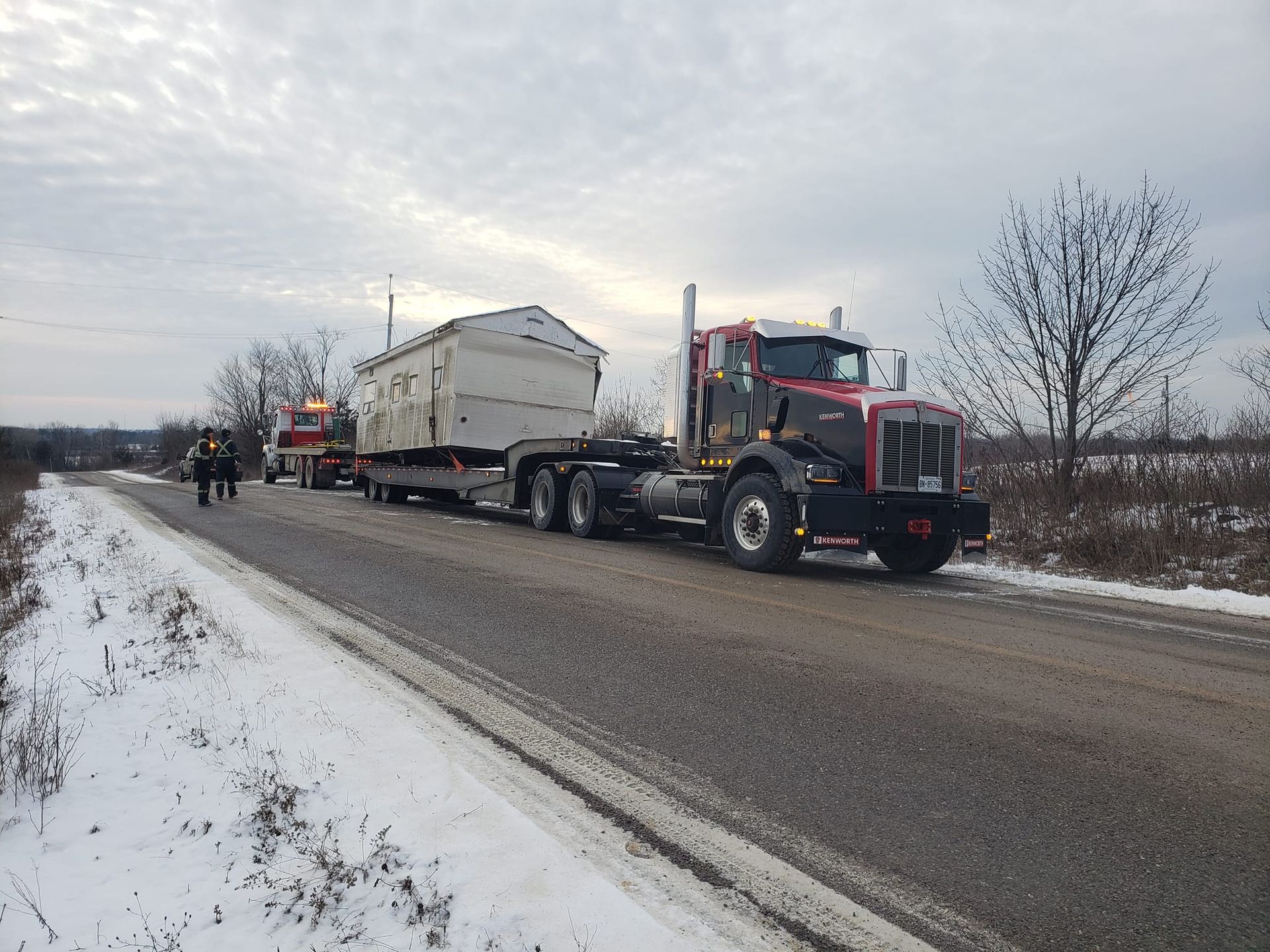 A semi-truck hauling a small building on a snowy roadside.