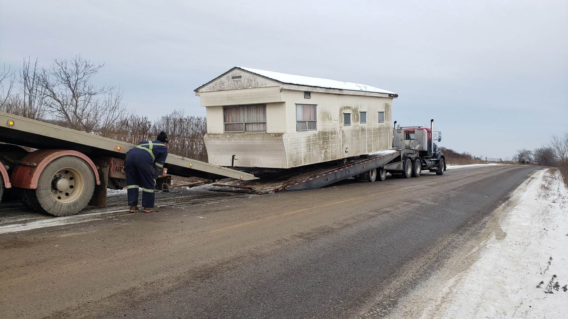 A mobile home being loaded onto a flatbed trailer on a snowy roadside. Two workers assist.