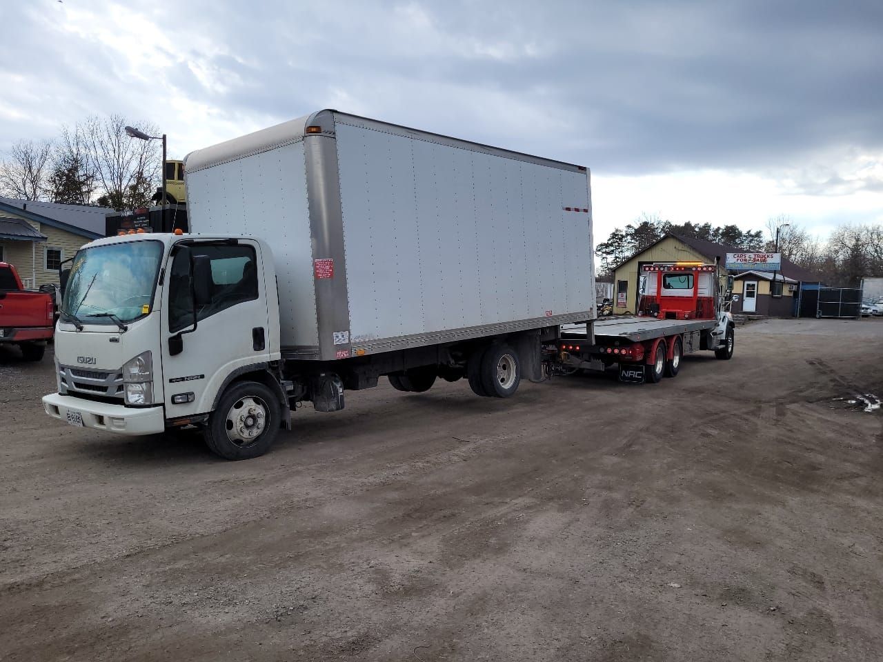 White box truck and red flatbed truck parked on gravel, cloudy sky.