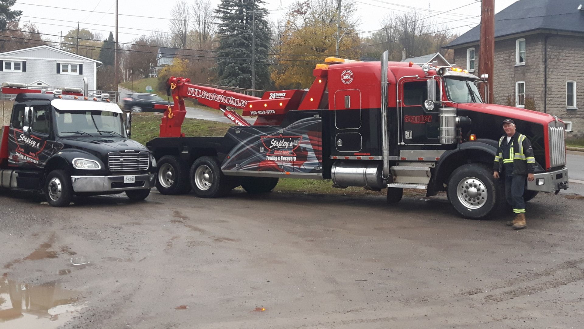 Two tow trucks, one black and one red, parked on a muddy lot, with a person in a safety vest.