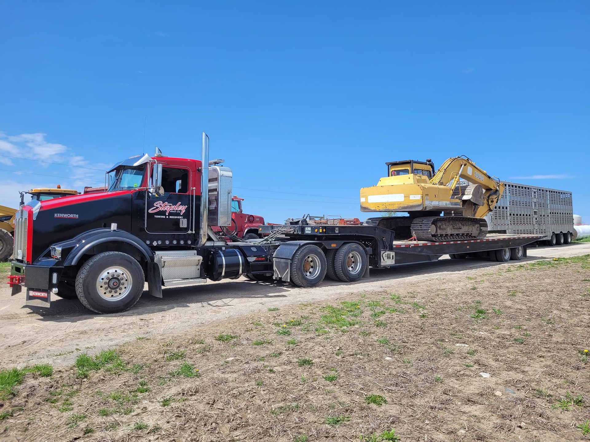 Black and red semi-truck with a flatbed trailer transporting a yellow excavator on a gravel road, blue sky.