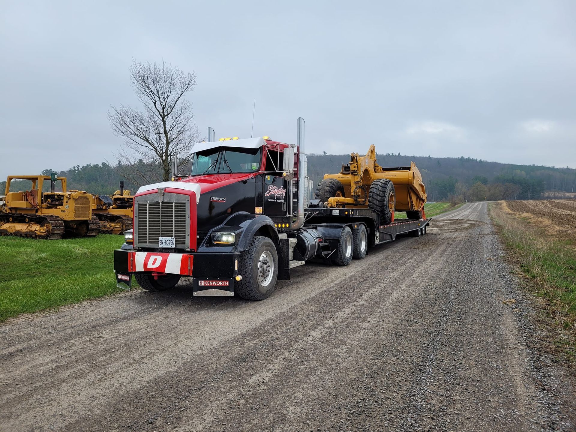 Black and red semi-truck transporting a yellow construction vehicle on a gravel road, overcast day.