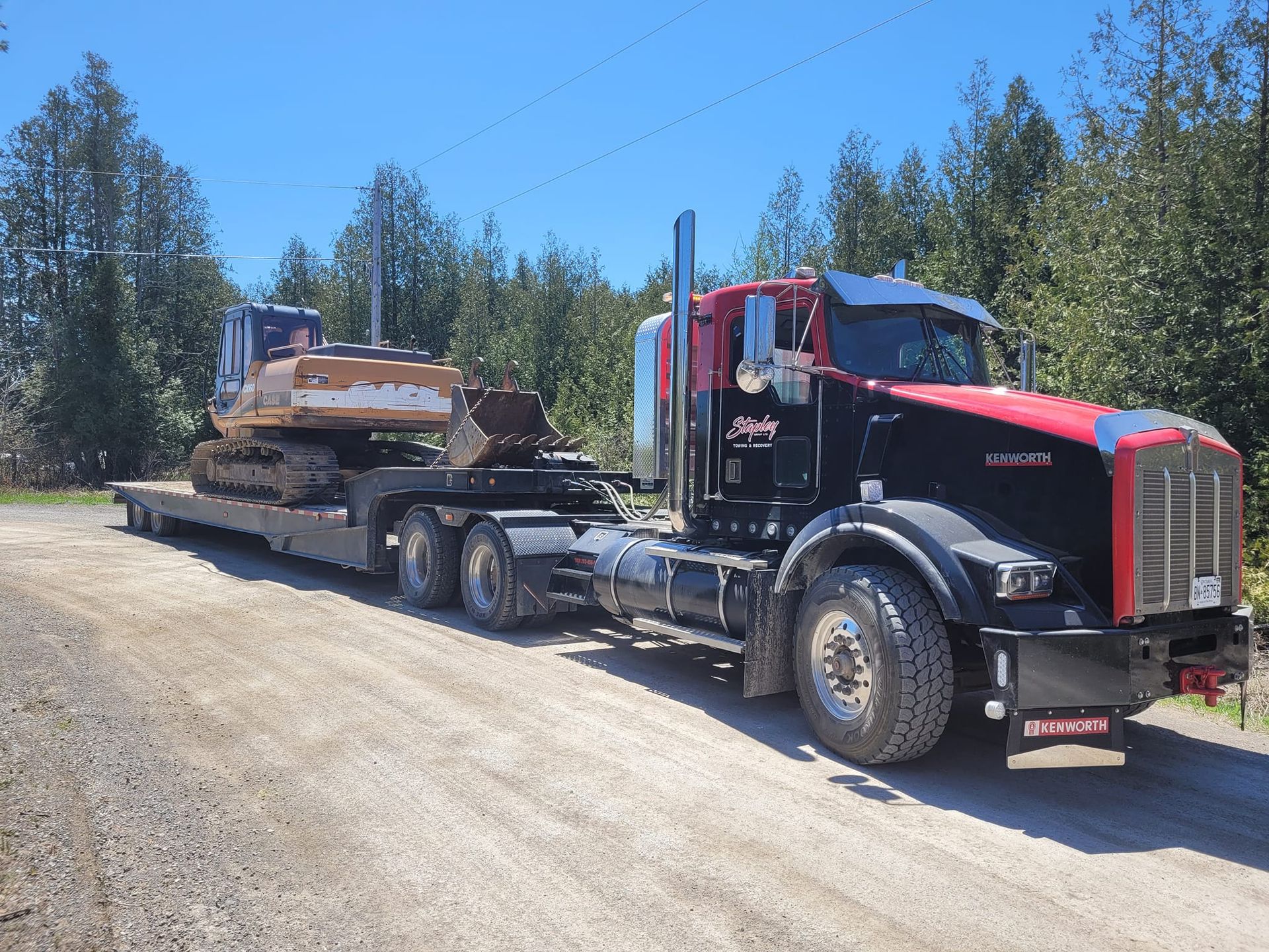 Black and red Kenworth truck with excavator on a lowboy trailer on a dirt road, trees in the background.