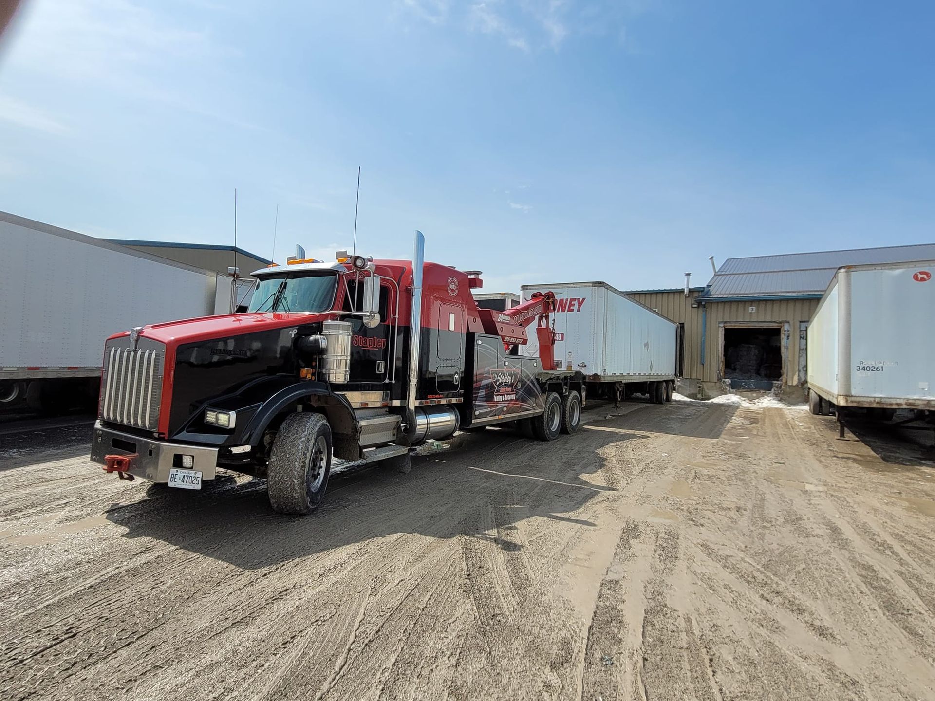 Red and black semi-truck towing a white trailer in a dirt lot. The setting is under a clear, bright blue sky.