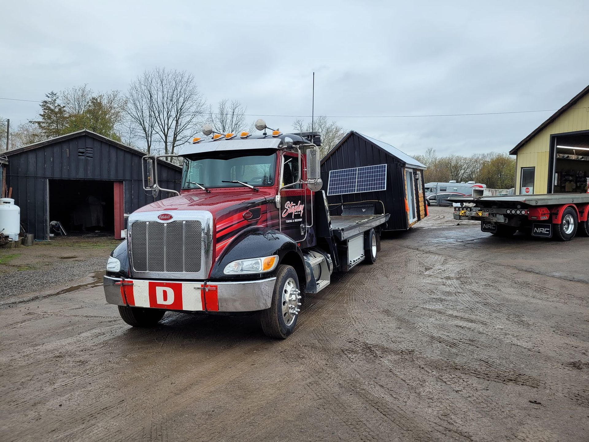 A flatbed tow truck hauling a small black cabin on a muddy lot, with a small shed and building in the background.