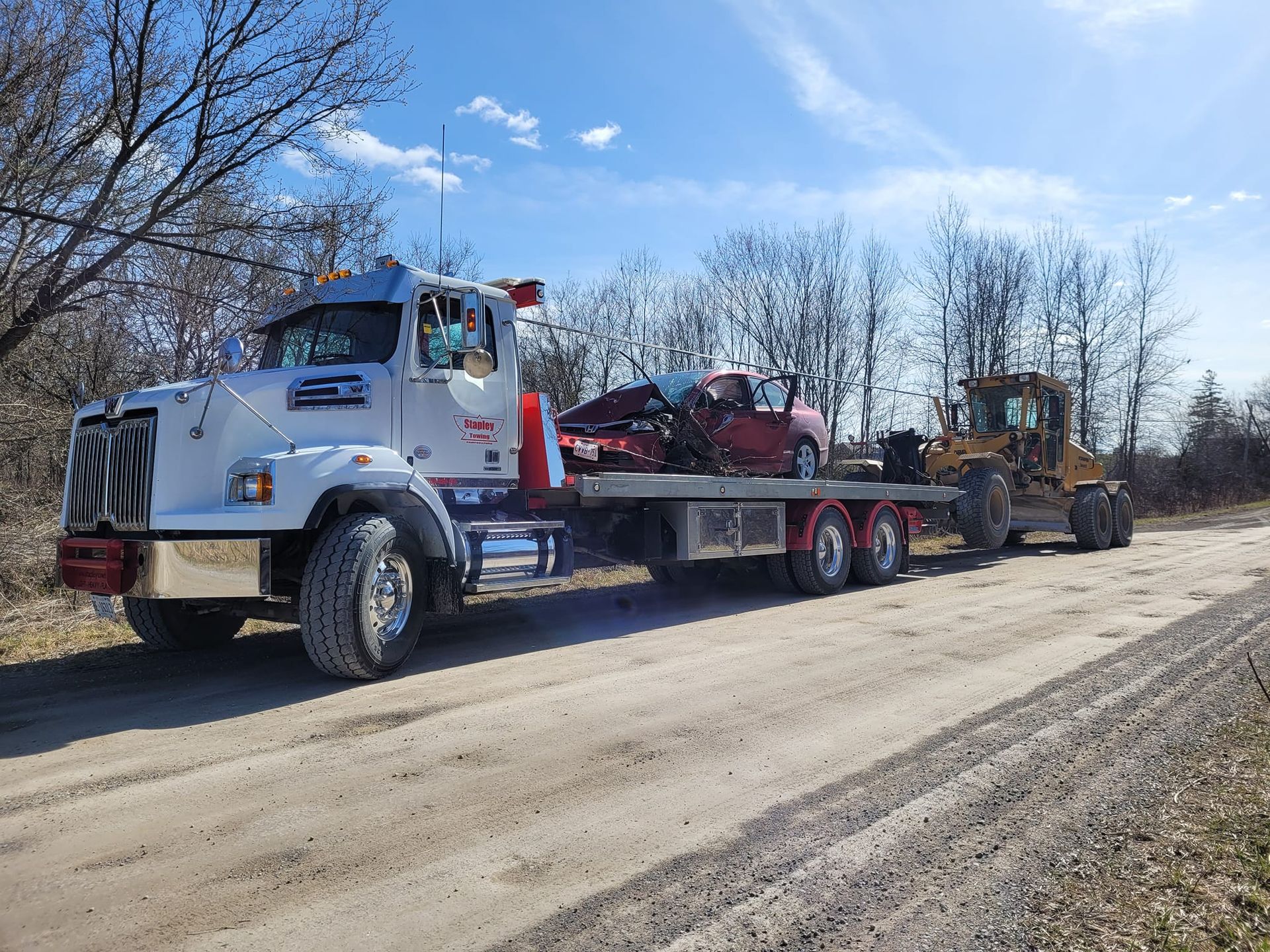 Tow truck carrying a wrecked red car and a piece of construction equipment on a dirt road under a sunny sky.