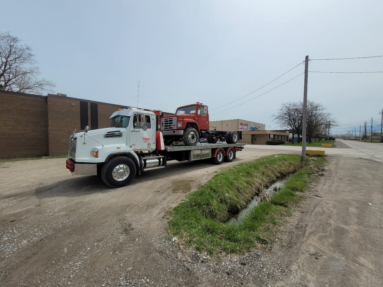 A white tow truck carries a red truck on a gravel road next to a ditch.