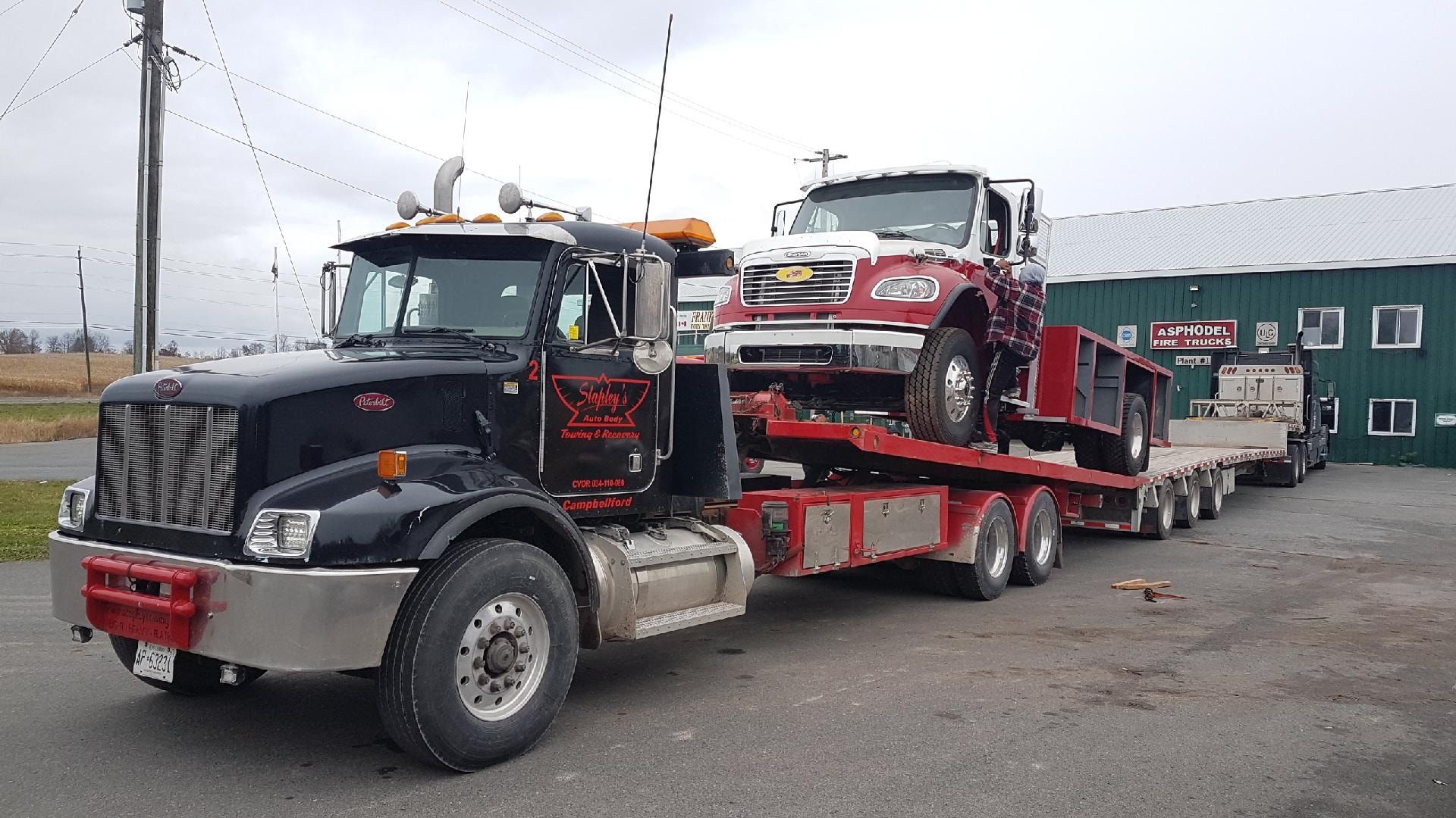 Black semi-truck hauling a red truck on a flatbed trailer in front of a green building.