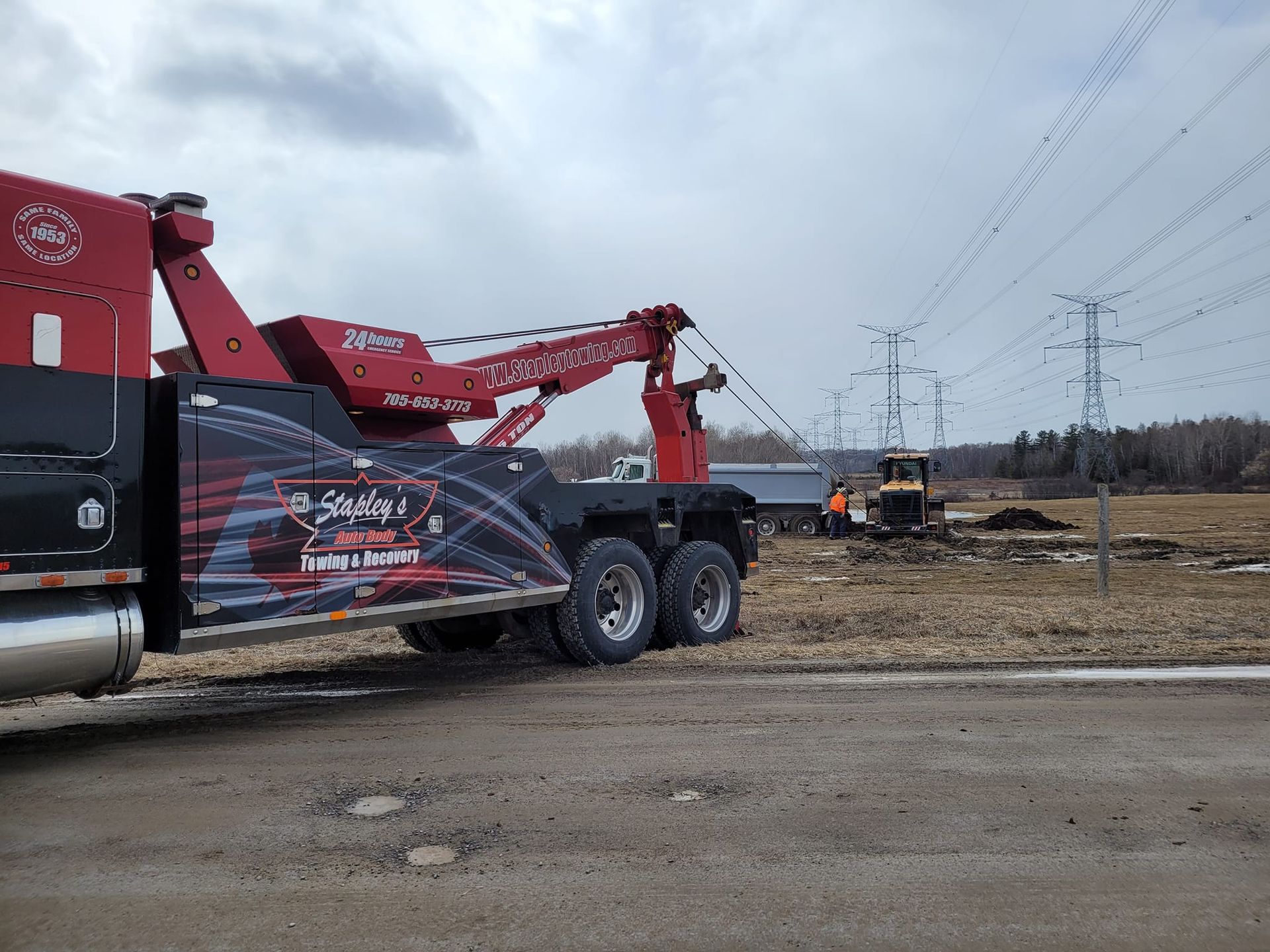 Tow truck lifting a small vehicle near power lines in a field on a cloudy day.