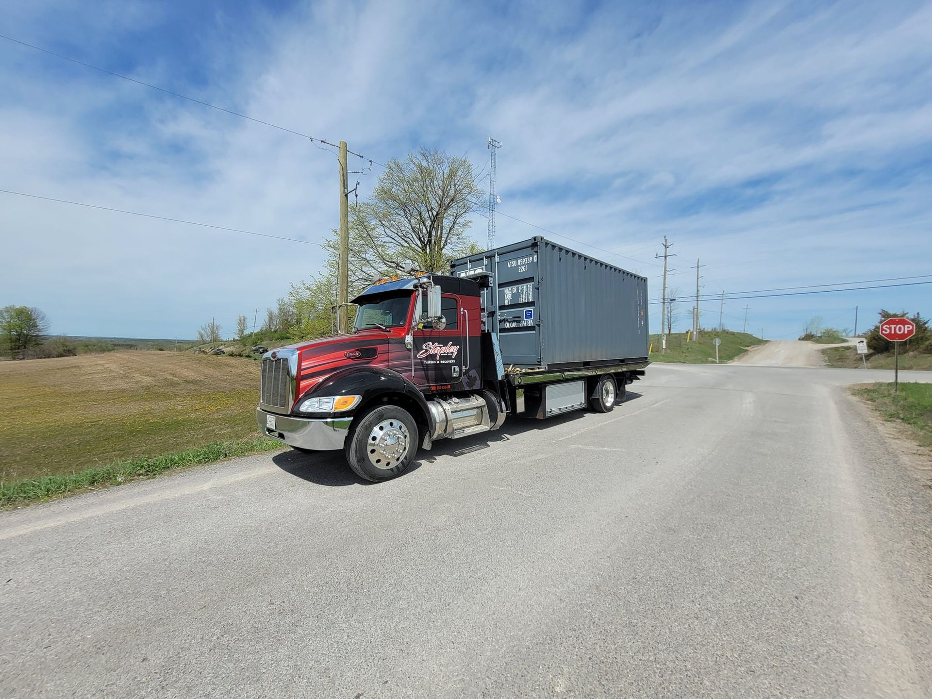Red semi-truck hauling a gray shipping container on a gravel road, with a blue sky and fields in the background.