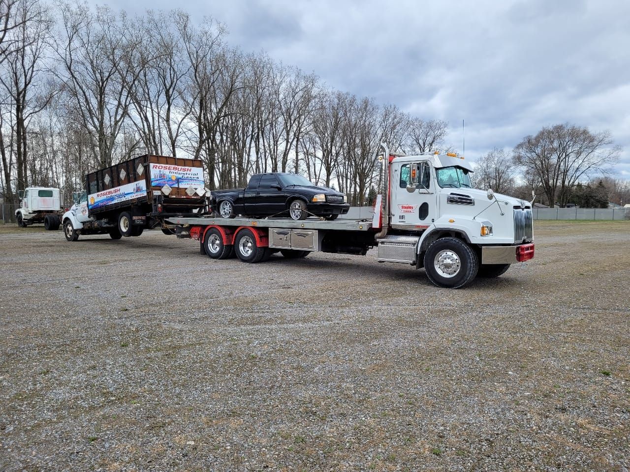 White tow truck hauling a black car on a flatbed trailer and a dumpster trailer on gravel, under a cloudy sky.