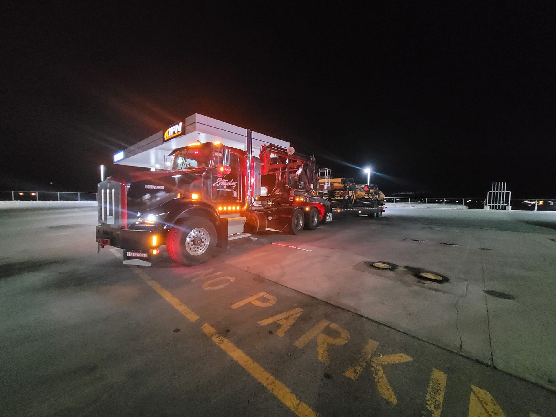 Dark semi-truck on a parking deck at night, hauling machinery, with illuminated headlights.
