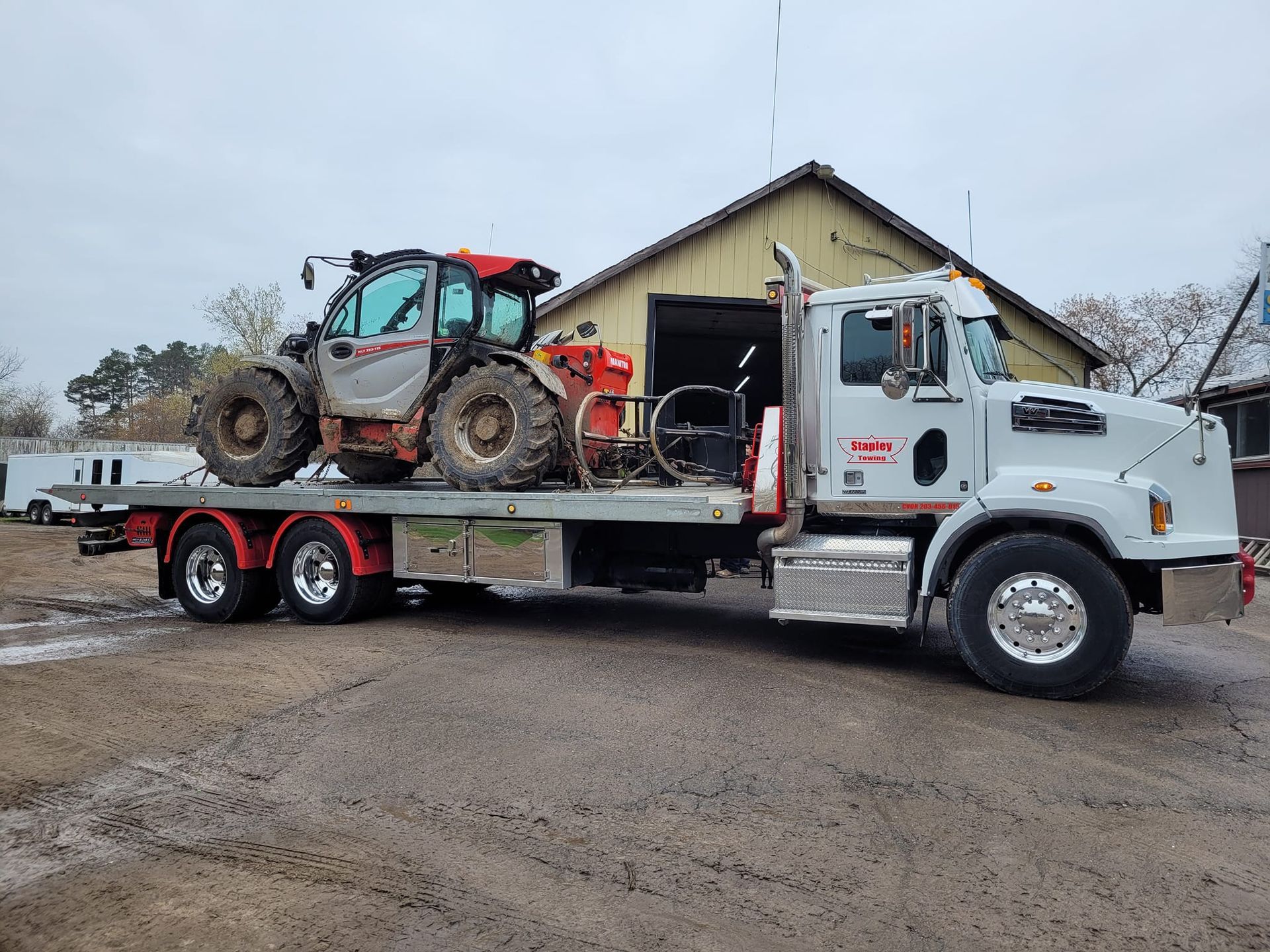 White tow truck carrying a red and gray telehandler on its flatbed in front of a barn.
