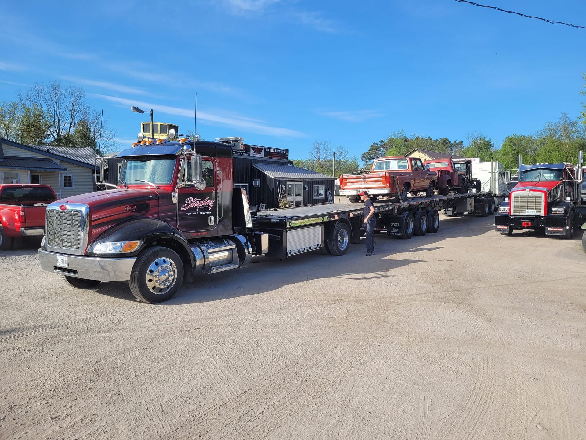 A large red and black tow truck carrying two classic orange pickup trucks on a flatbed. Sunny day.