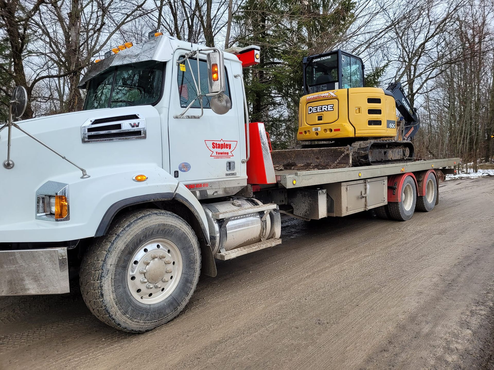 White truck with a yellow excavator on a flatbed trailer, on a dirt road.