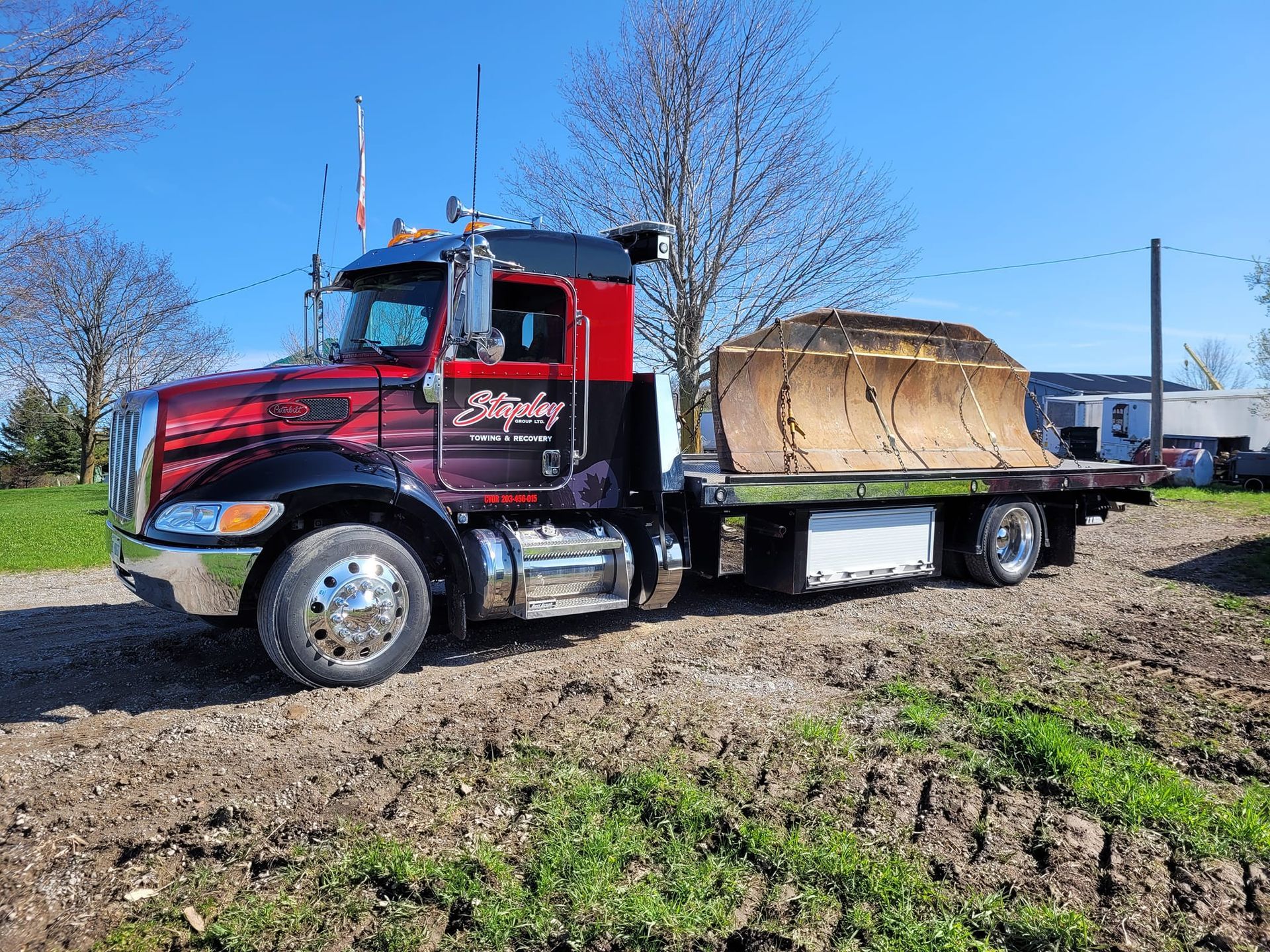 Red and black semi-truck hauling a large, covered object on a flatbed trailer, parked on dirt.
