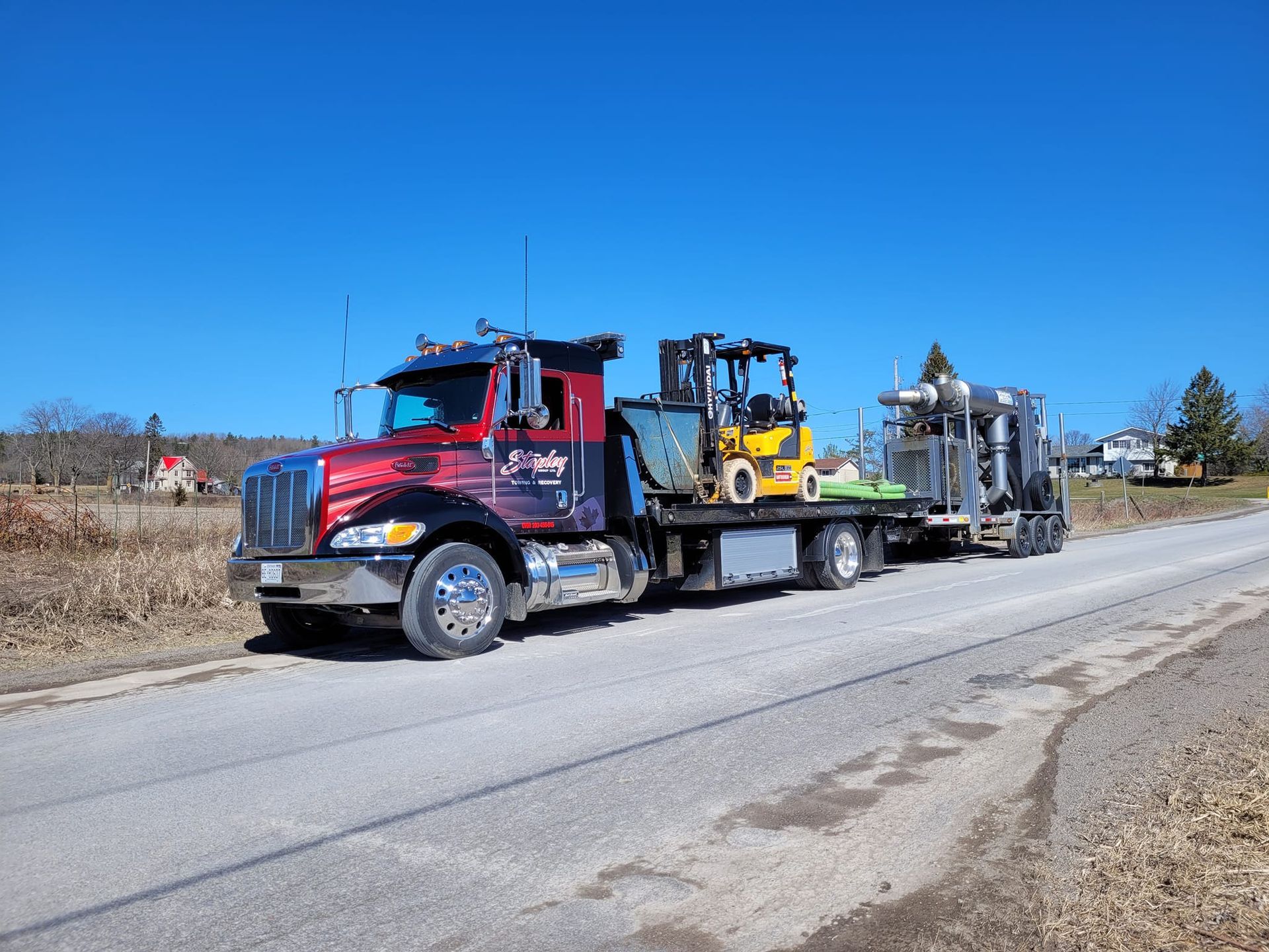 Red semi-truck hauling a yellow and blue forklift on a flatbed trailer on a paved road under a blue sky.