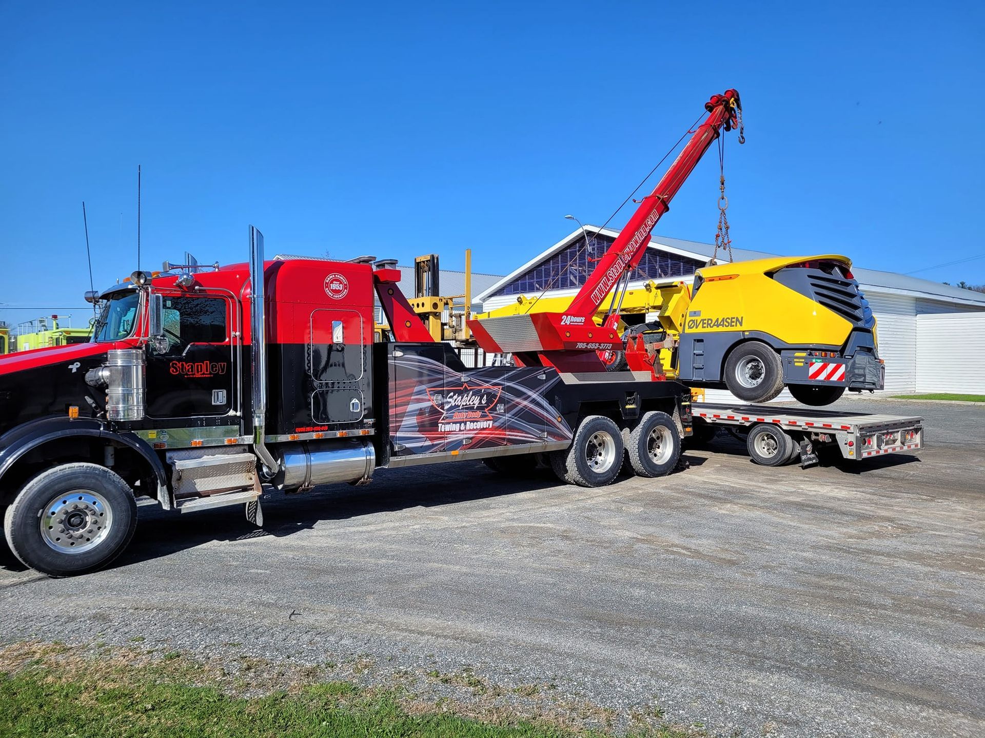 Red tow truck loading yellow and gray industrial equipment onto a trailer, outdoors on a sunny day.
