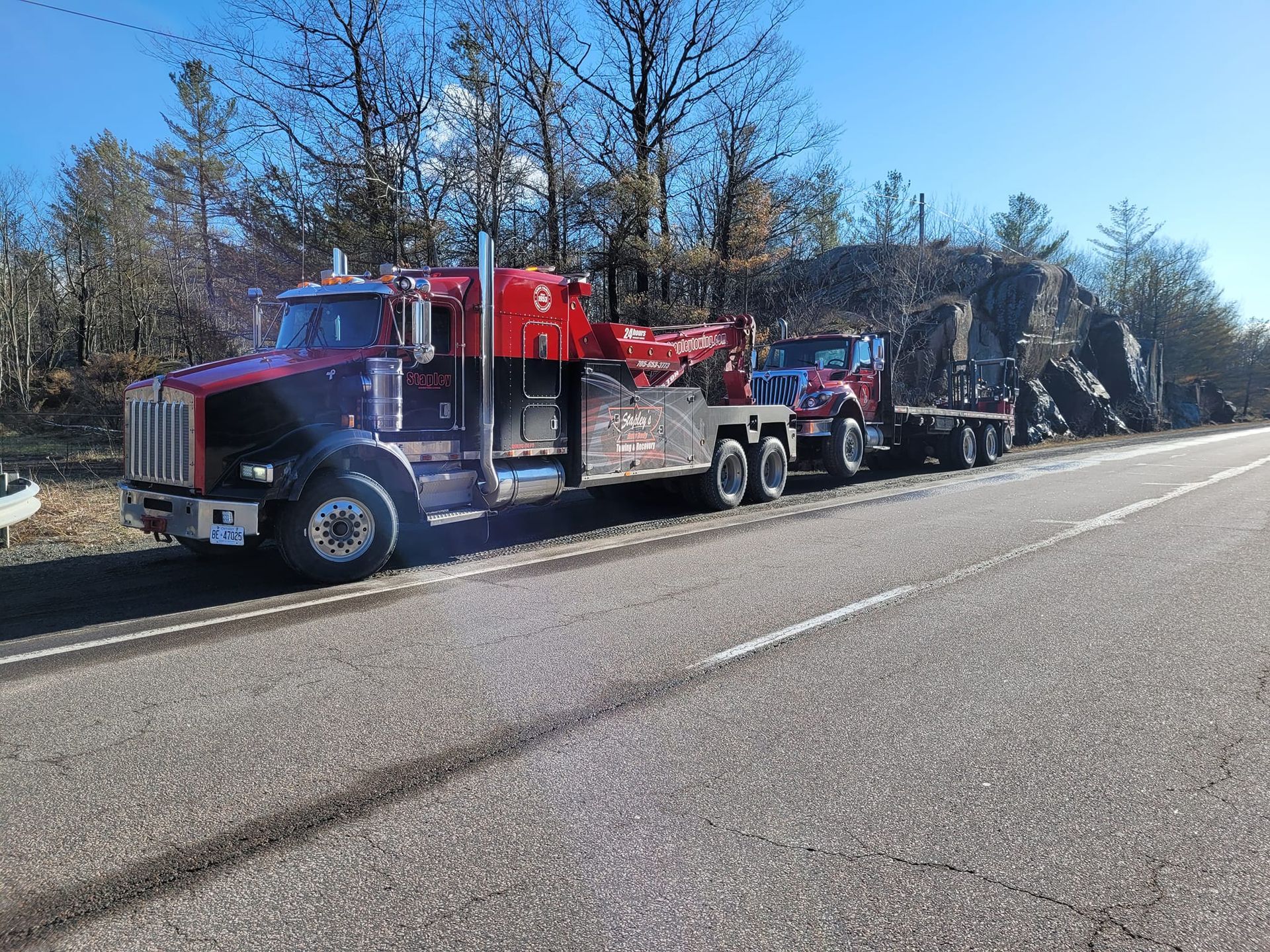 Red semi-truck with a flatbed trailer carrying a smaller red truck on a roadside, under a bright sky.