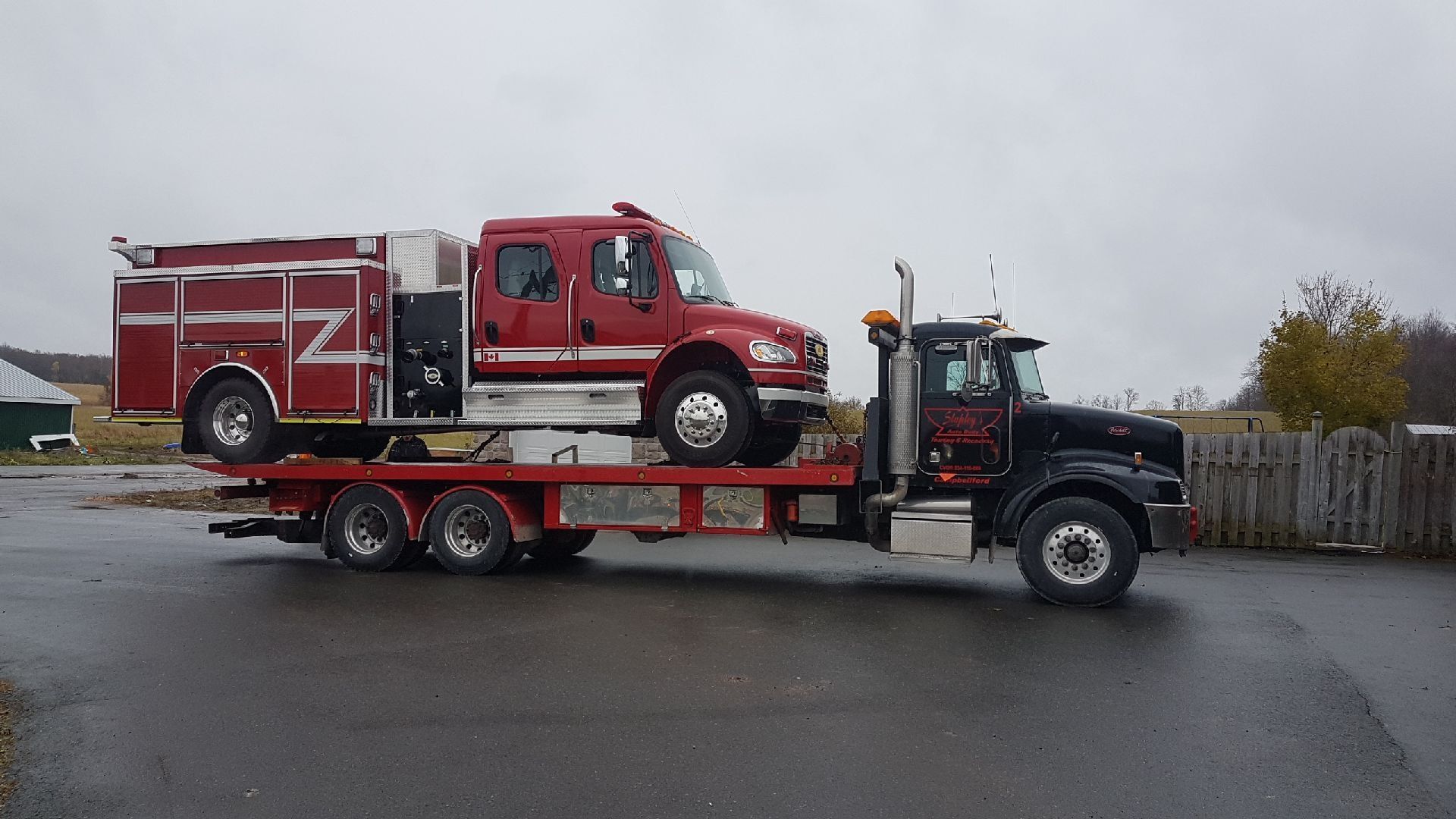 Red fire truck on a flatbed tow truck, parked outdoors on a cloudy day.