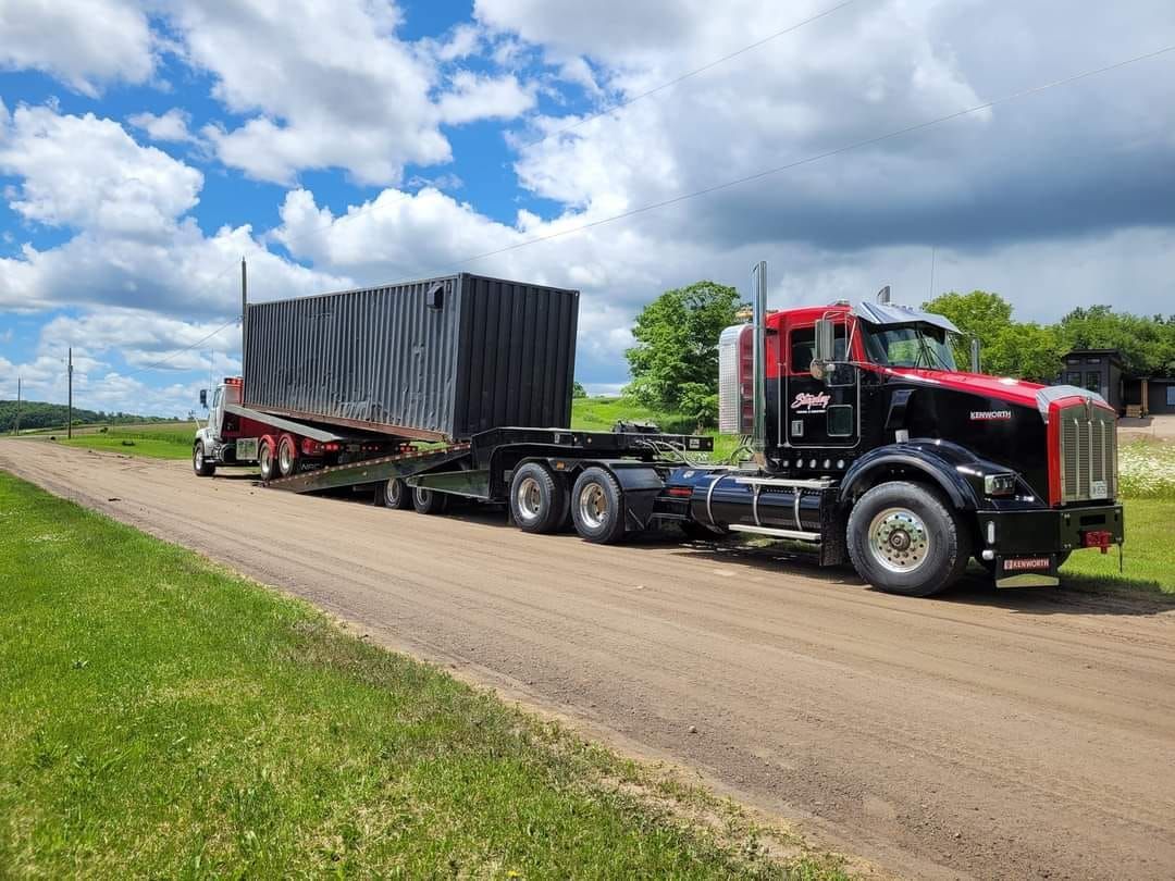 Black semi-truck hauling large, black container on a lowboy trailer on a dirt road. Bright sky, green grass.