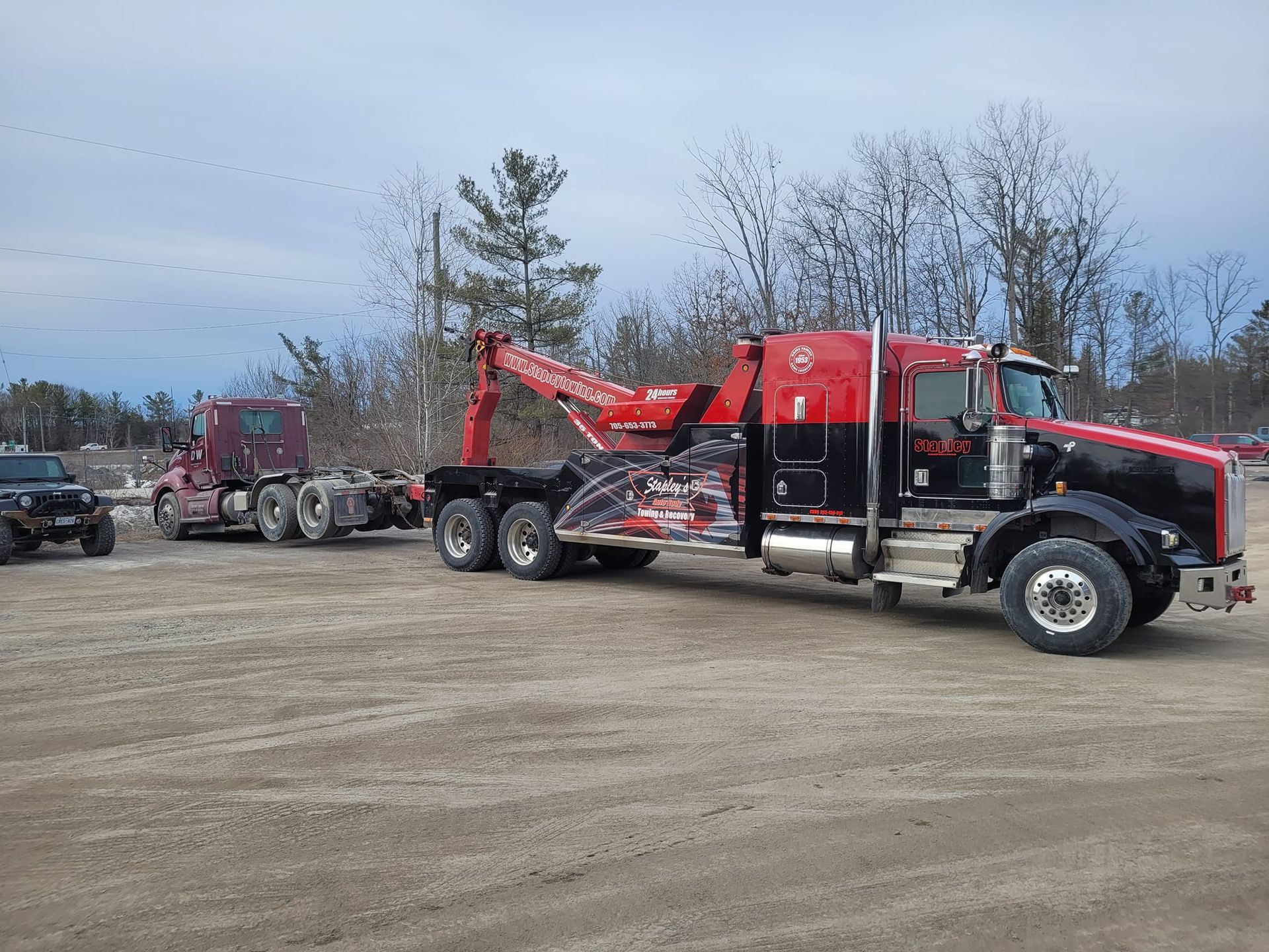 Tow truck towing a semi-truck on a gravel lot. Red and black truck, cloudy sky, trees in the background.