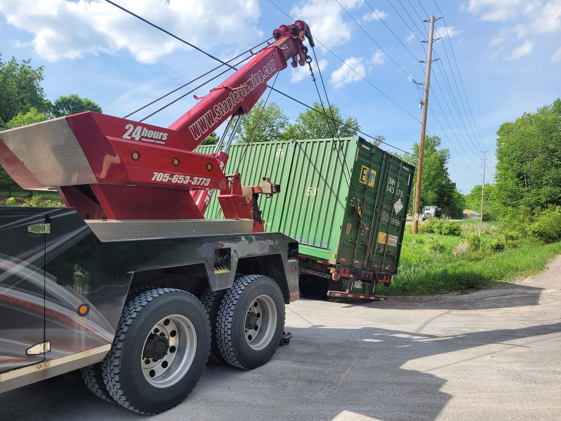 Tow truck lifting a green shipping container from a rural roadside. Power lines and trees in the background.