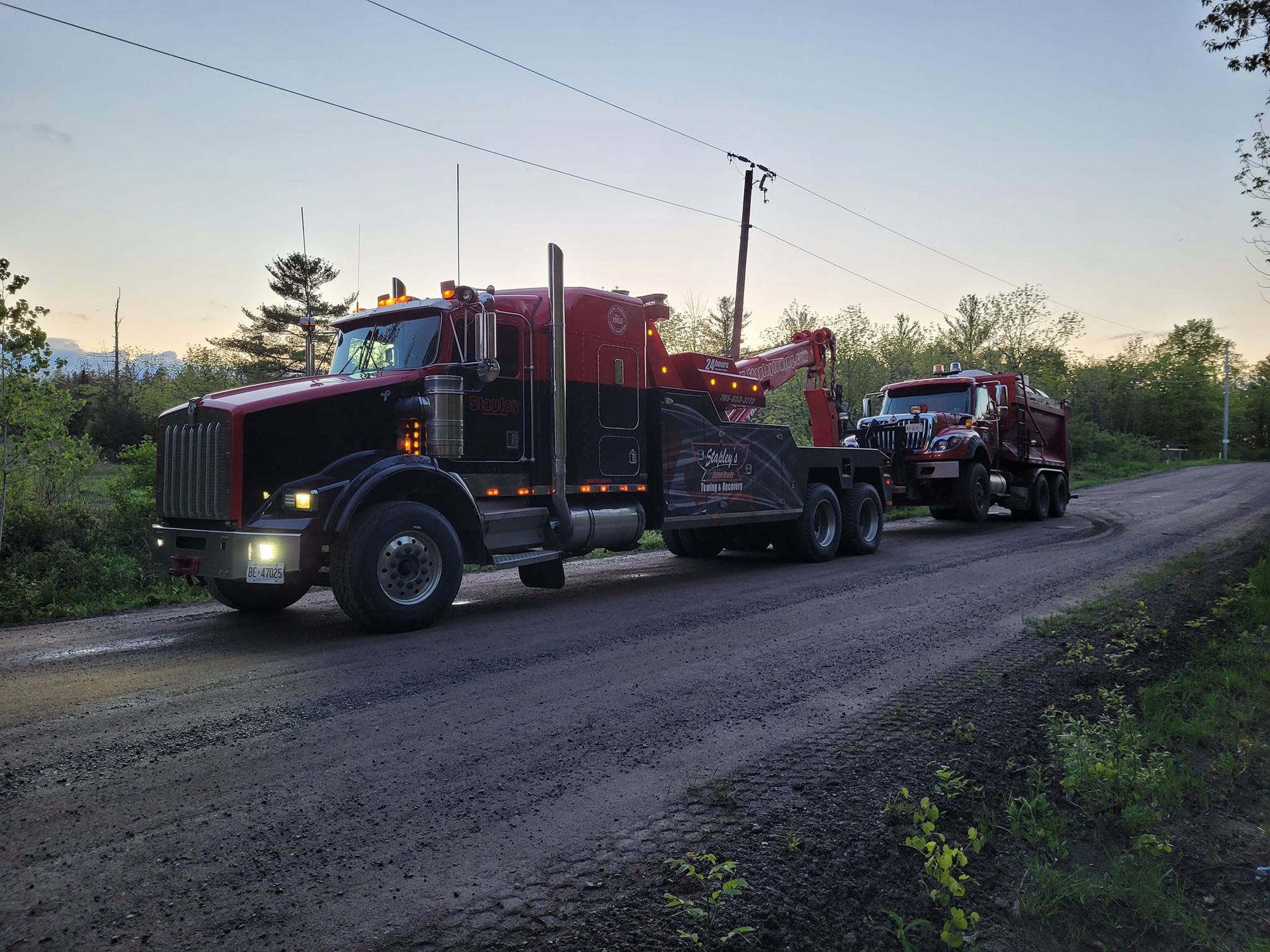 Red and black tow truck pulling a red truck on a dirt road at dusk.