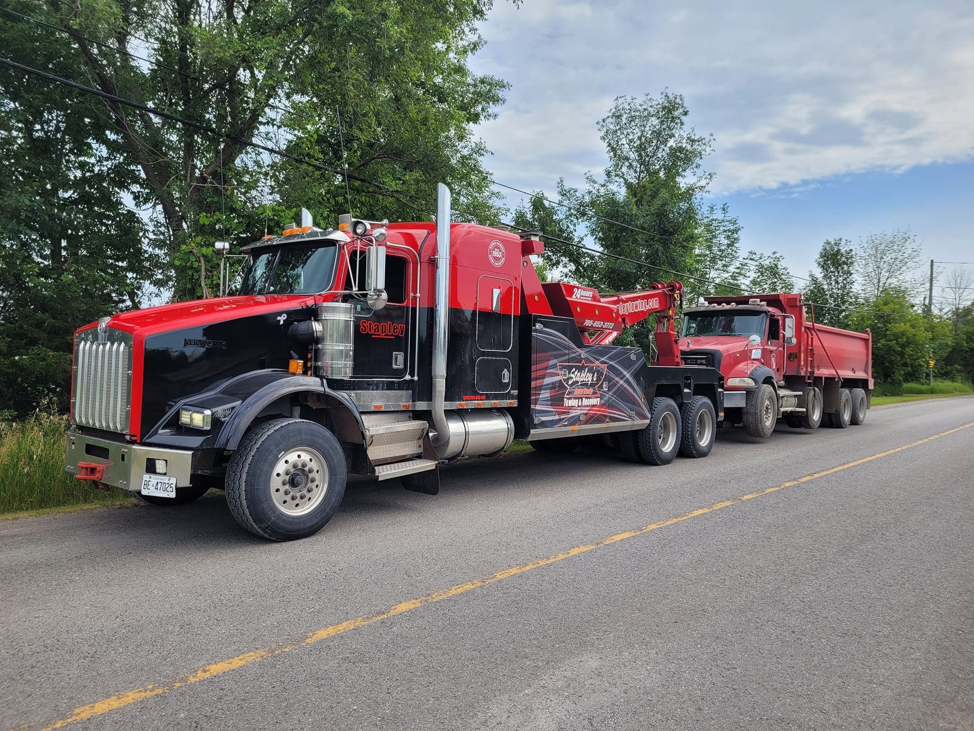Red and black tow truck towing a red dump truck on a road.