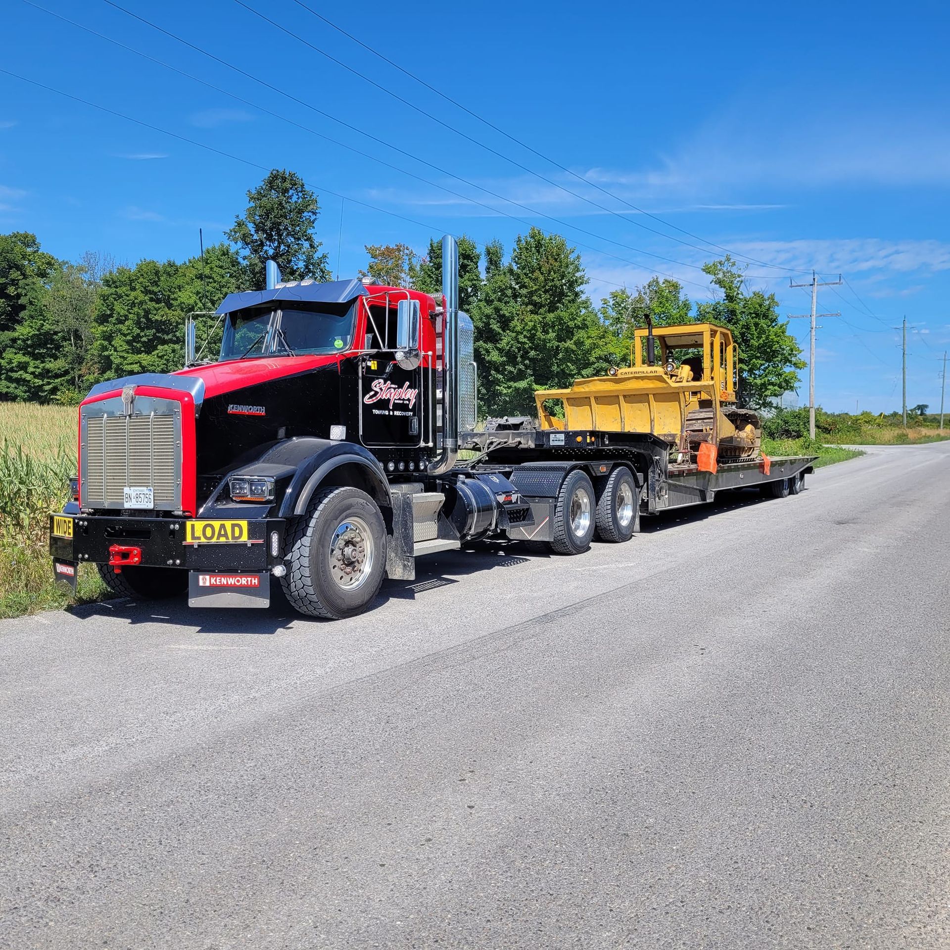 Black and red semi-truck hauling a yellow bulldozer on a flatbed trailer on a rural road.