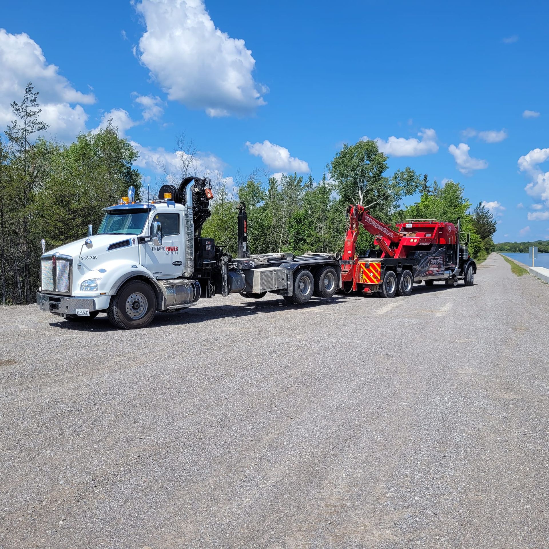 White truck with crane towing a red tow truck on a gravel road, blue sky, trees.