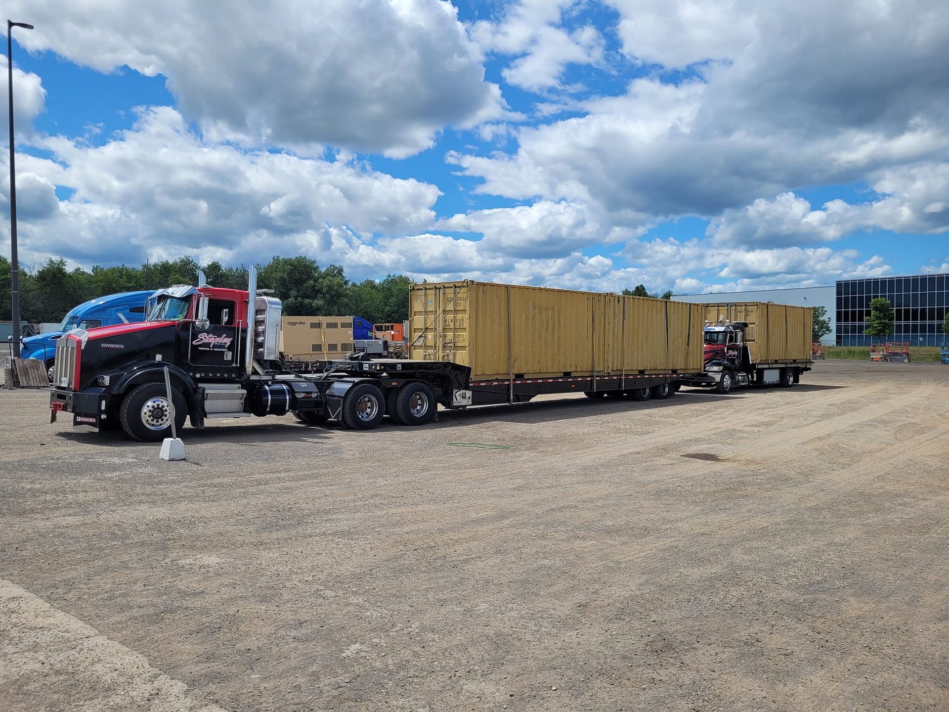 Two semi-trucks parked on gravel, each hauling long, beige shipping containers under a cloudy blue sky.
