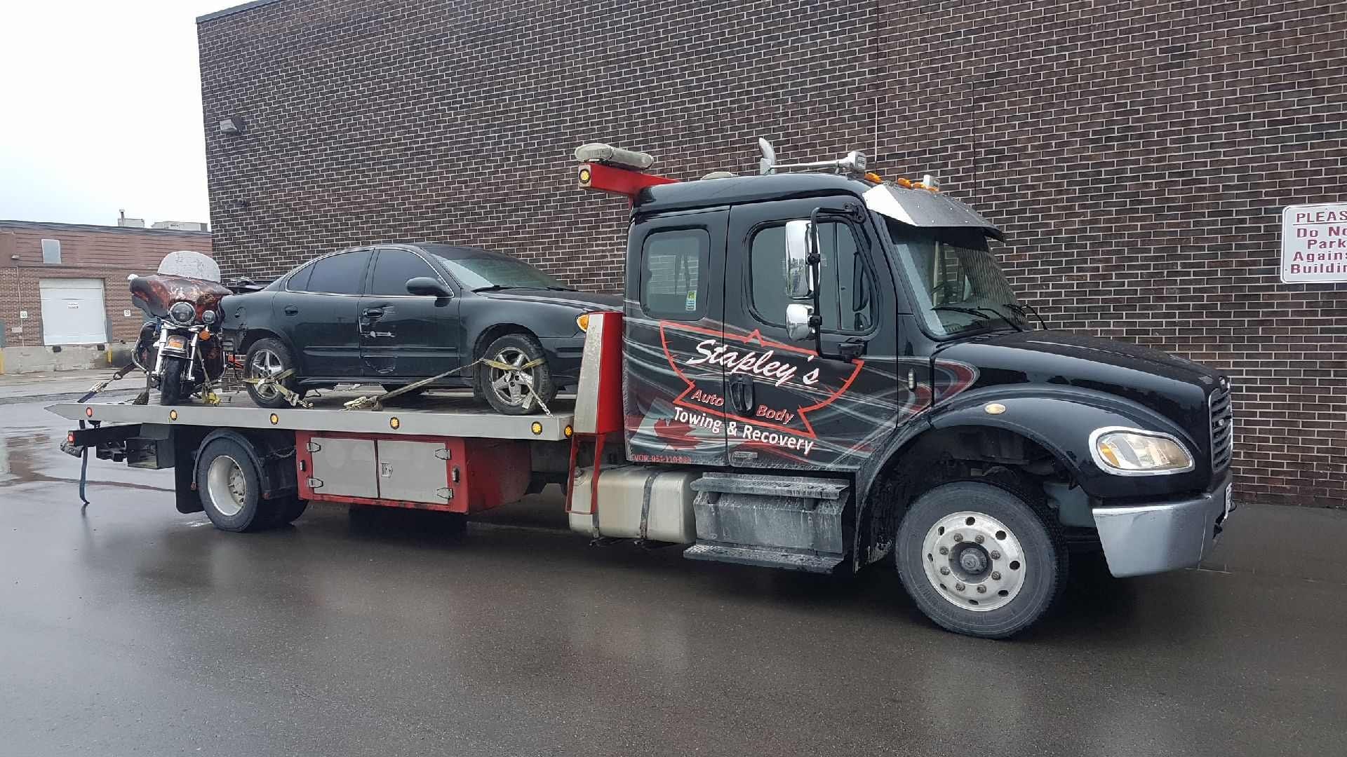 A black tow truck with a black car on the flatbed, parked in front of a brick building.