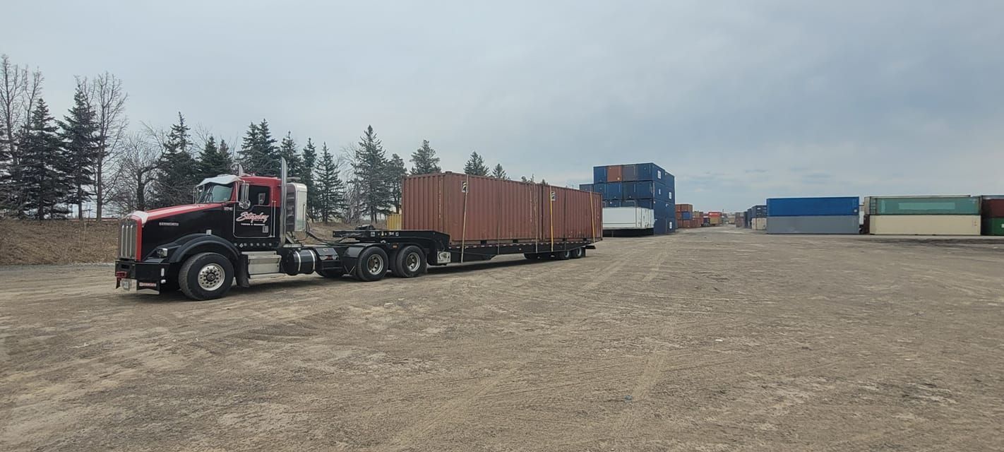 A black semi-truck hauling shipping containers in a storage yard under a cloudy sky.