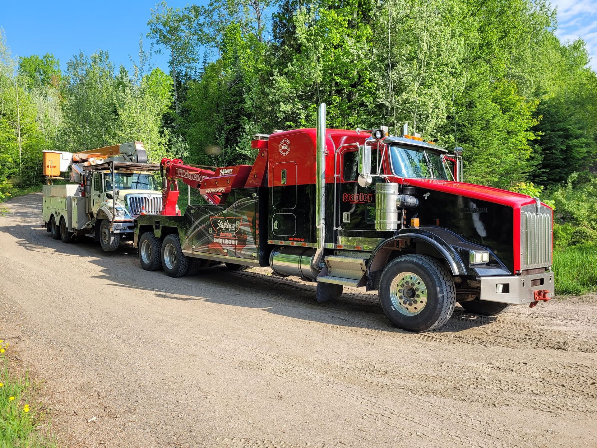 Red and black tow truck towing a white truck on a gravel road, trees in the background.