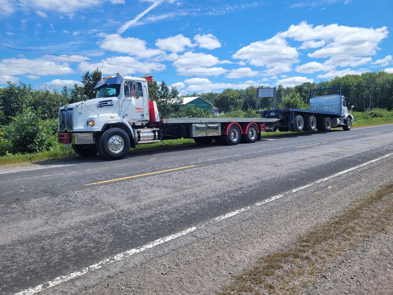 White flatbed truck on the side of a road, pulling a trailer with a blue container. Bright sunny day.