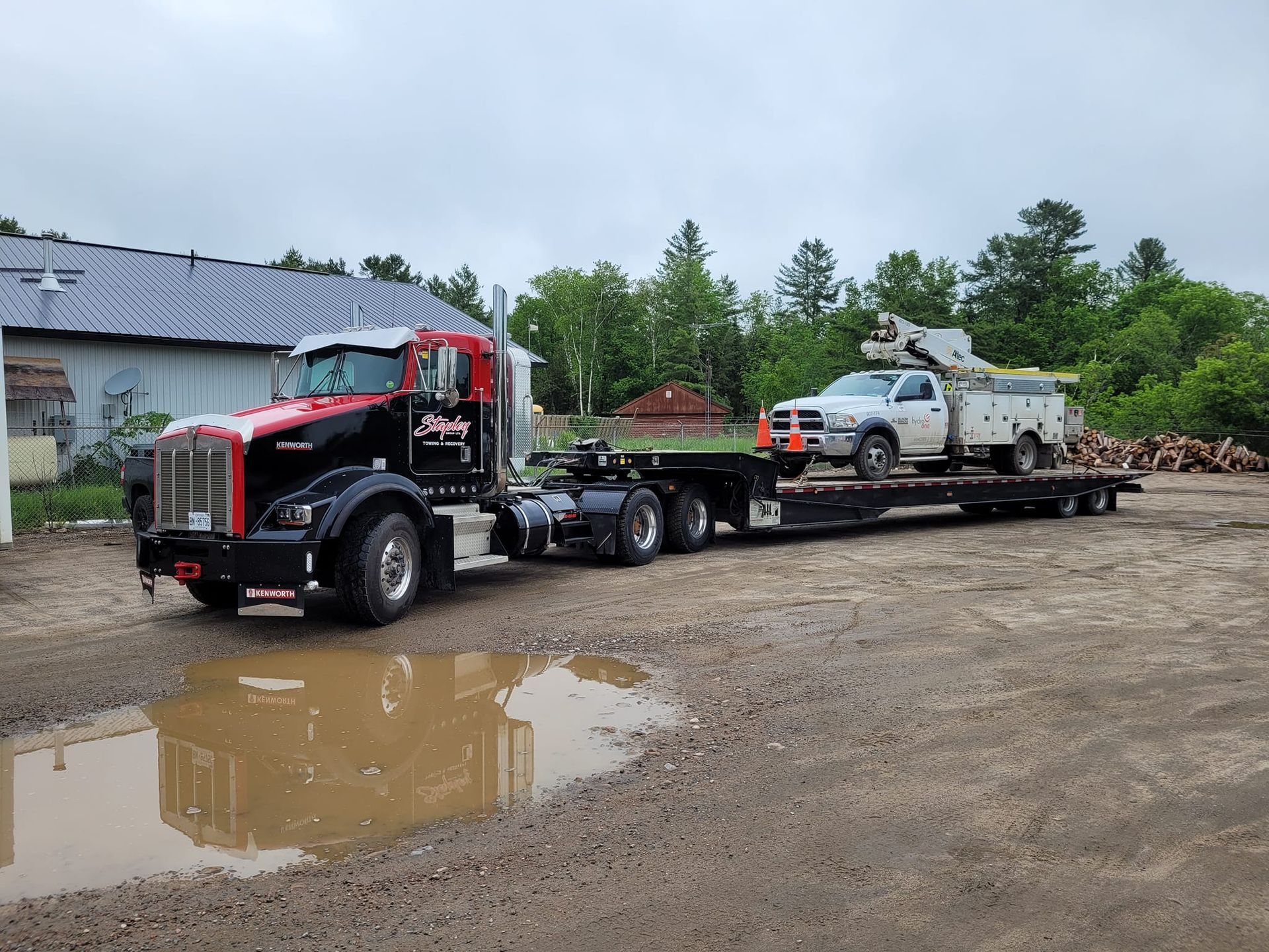 Black and red semi-truck hauling a white service truck on a flatbed trailer, parked on gravel next to a puddle.