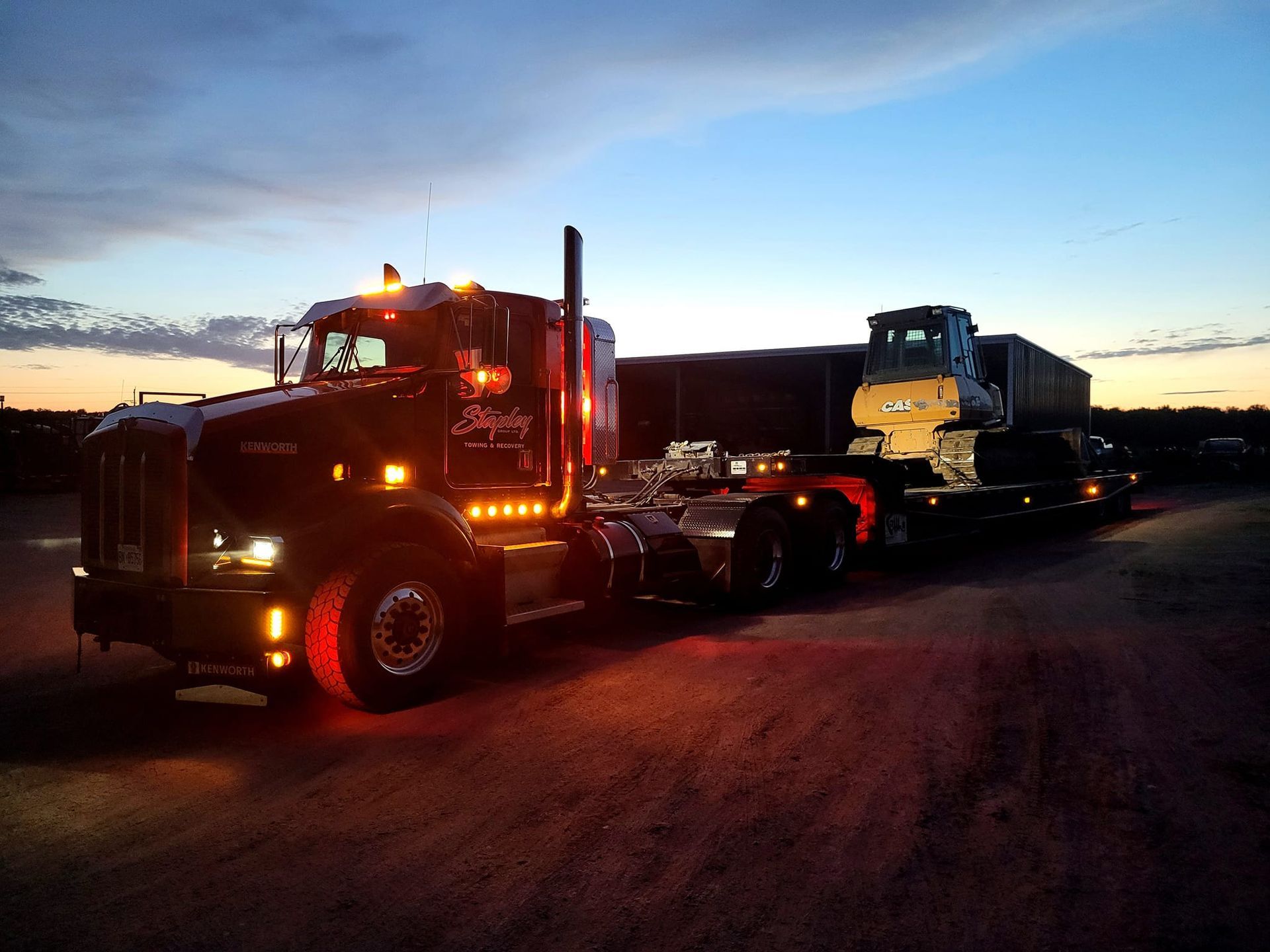 Semi-truck with a lowboy trailer carrying construction equipment at dusk.