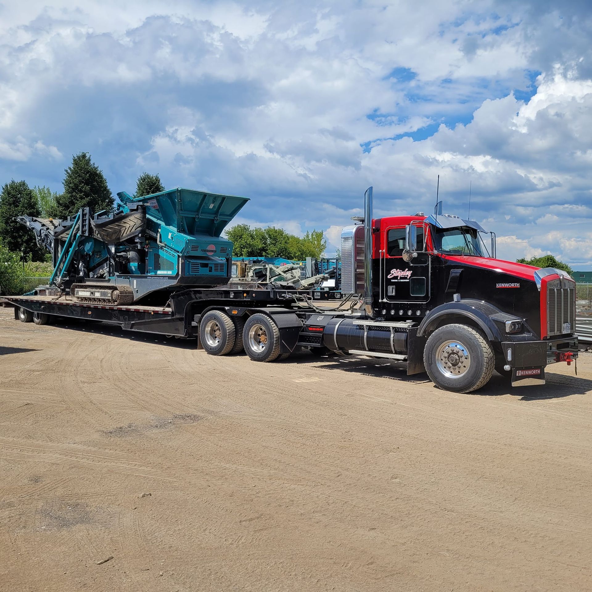 Black and red semi-truck hauling a teal industrial machine on a flatbed trailer, set on a gravel lot under a cloudy sky.