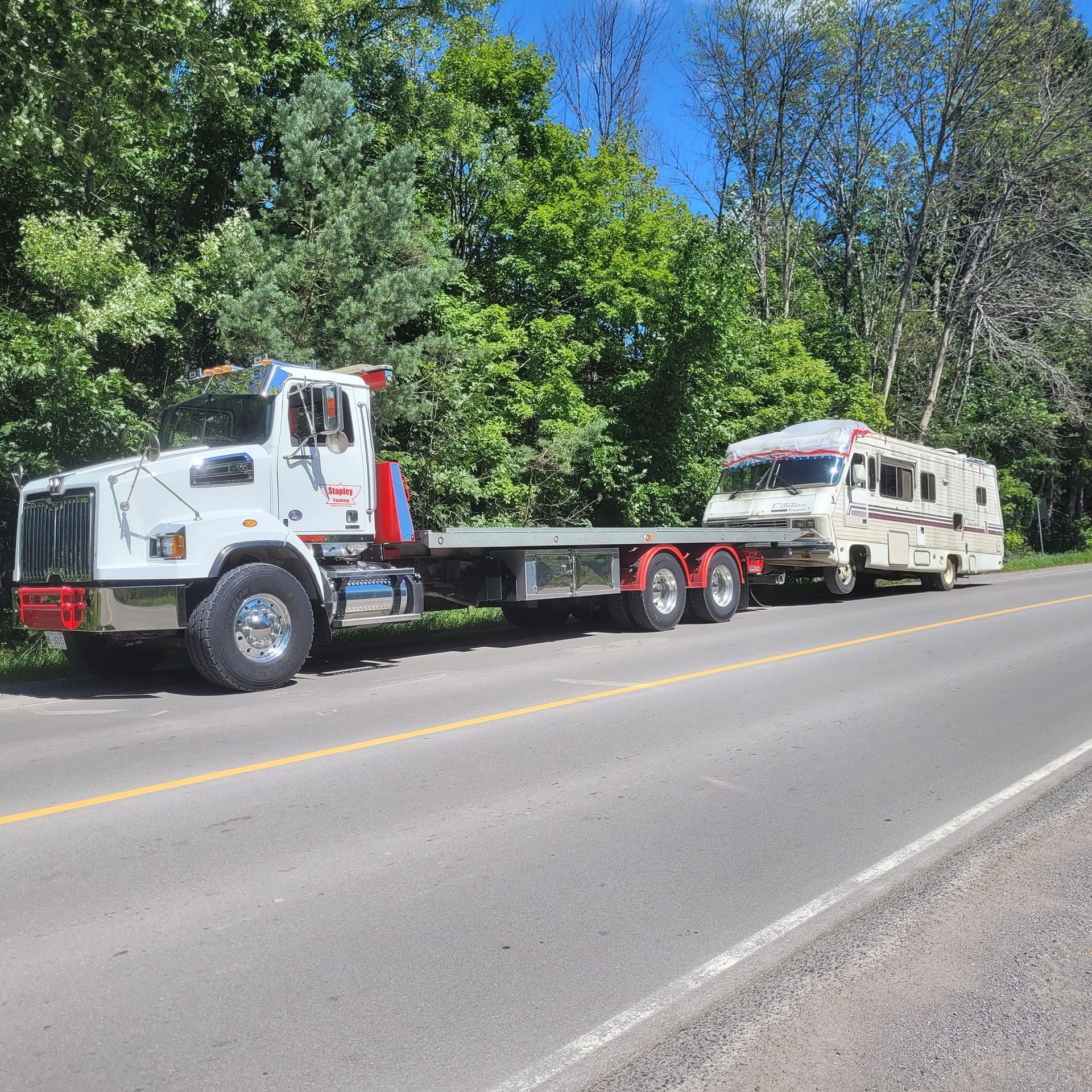 White tow truck hauling a tan RV on a flatbed along a road, trees in the background, sunny day.