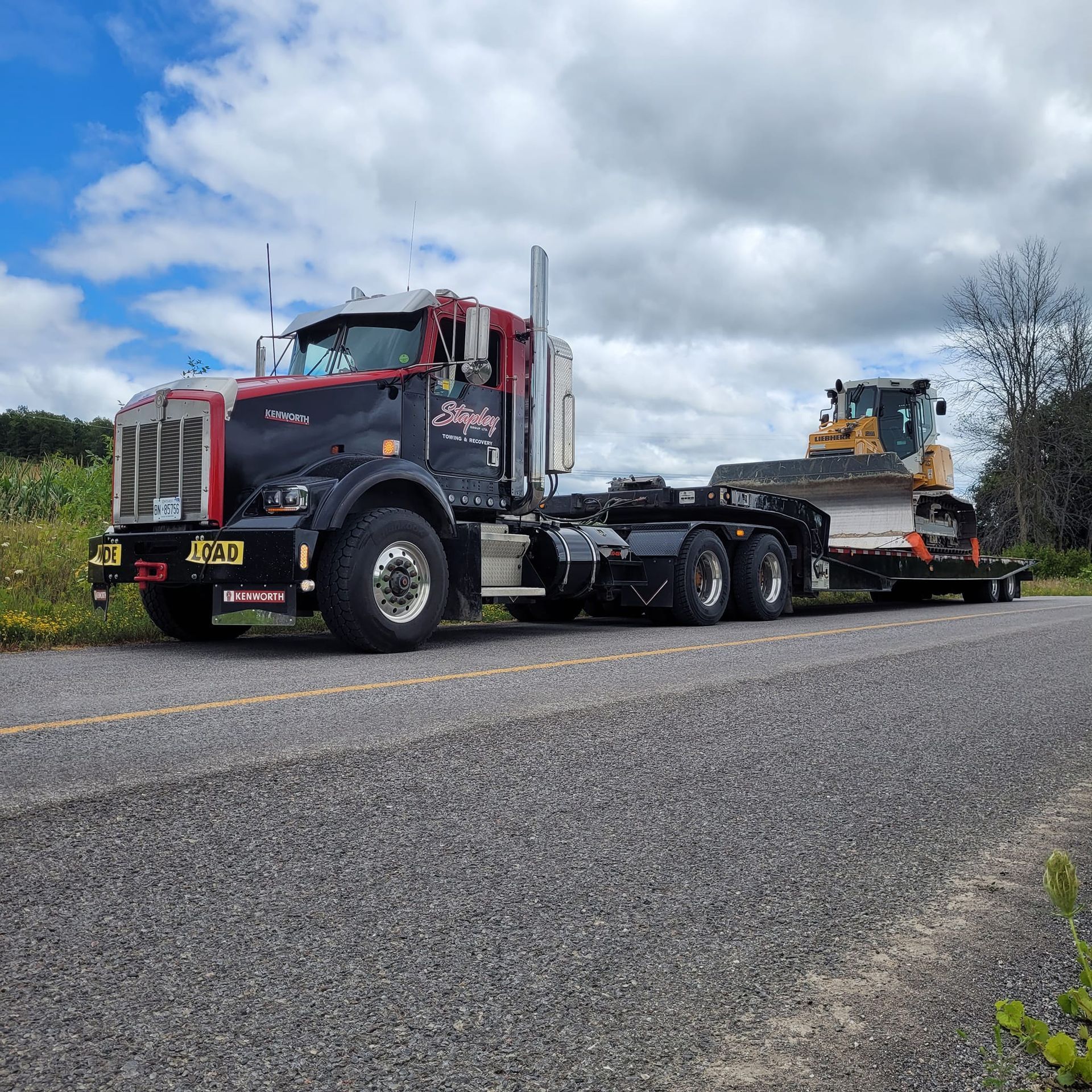 Black and red semi-truck with a flatbed trailer transporting a yellow bulldozer on a road, under a cloudy sky.