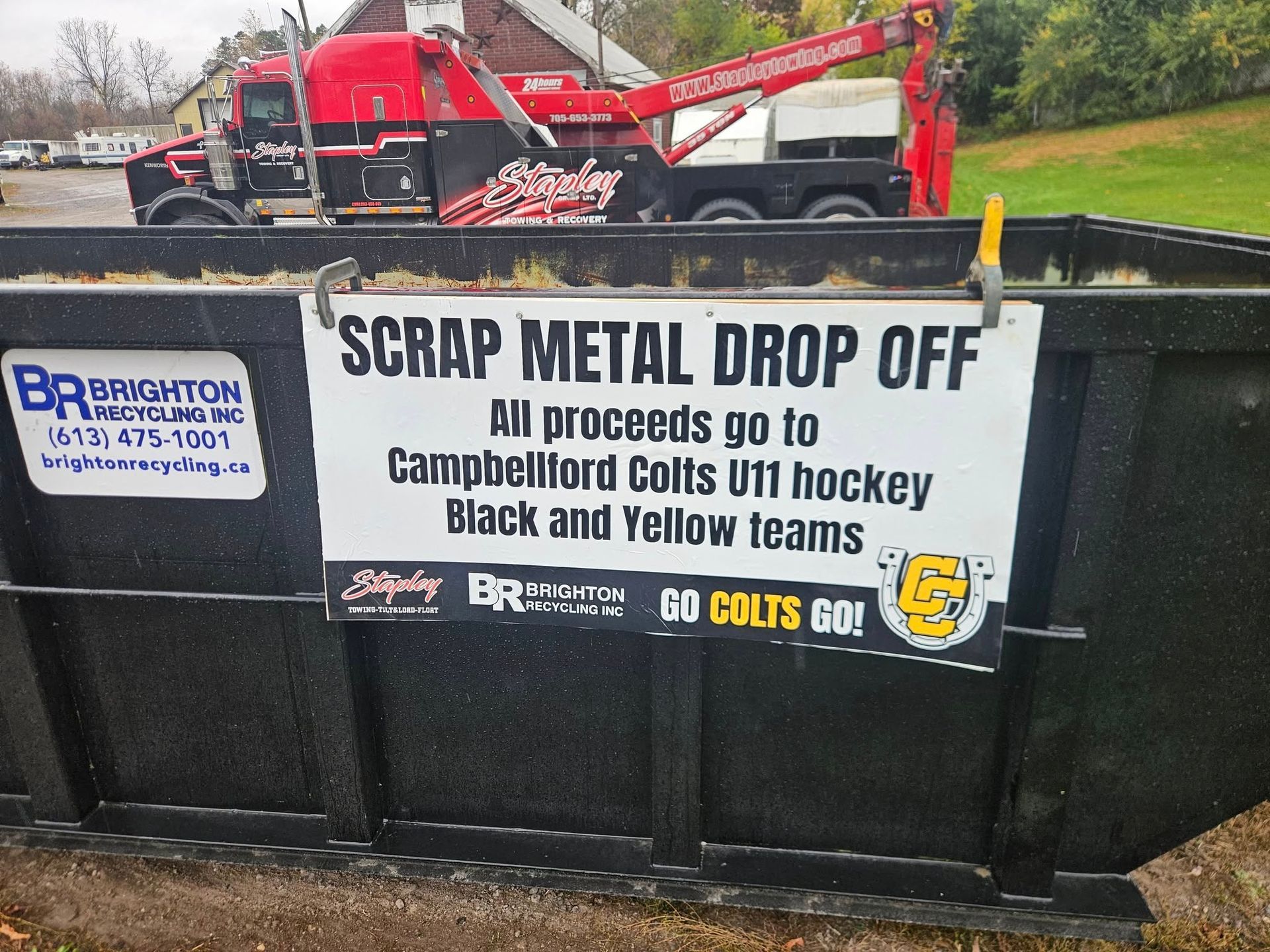 Scrap metal drop-off bin with sign for Campbellford Colts U11 hockey team. Red tow truck in background.